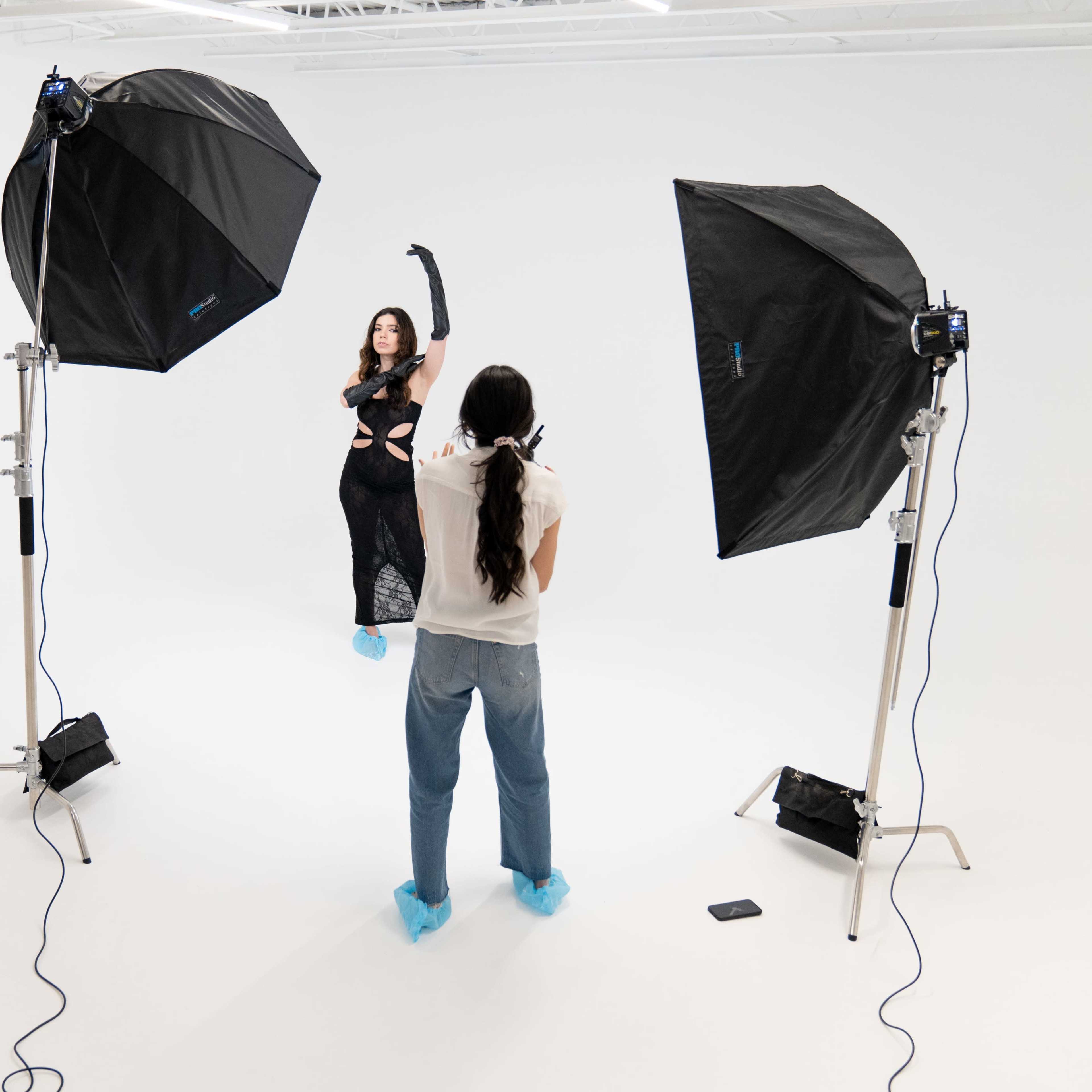 A model poses in a black outfit while a photographer captures the shot in a well-lit studio with two large softbox lights.