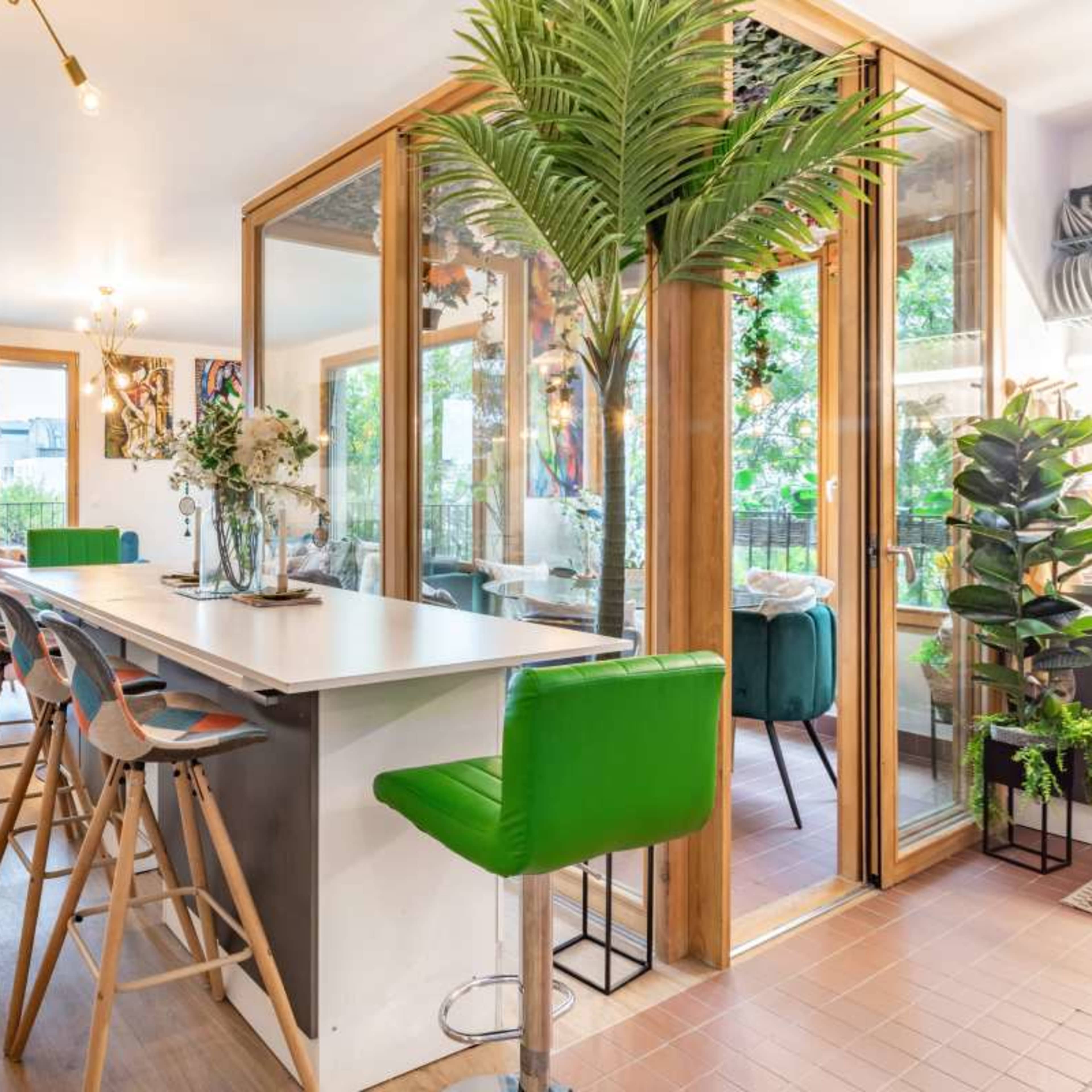 A spacious, modern kitchen and dining area features a long island with bar stools, large windows, and indoor plants for a vibrant atmosphere.