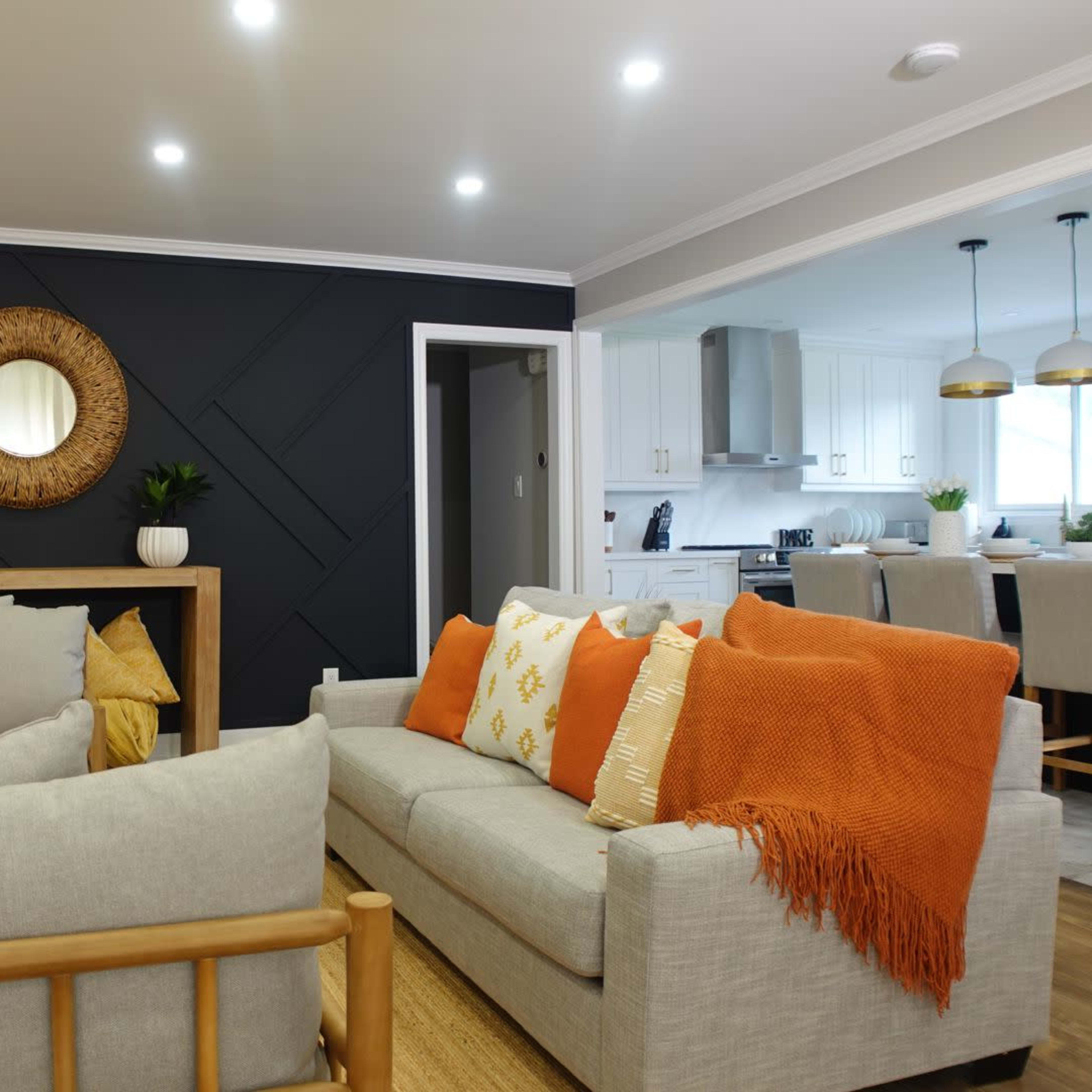 A living room features a light-colored sofa, an orange throw blanket, and a decorative wall with a round mirror, adjacent to a kitchen with white cabinetry.
