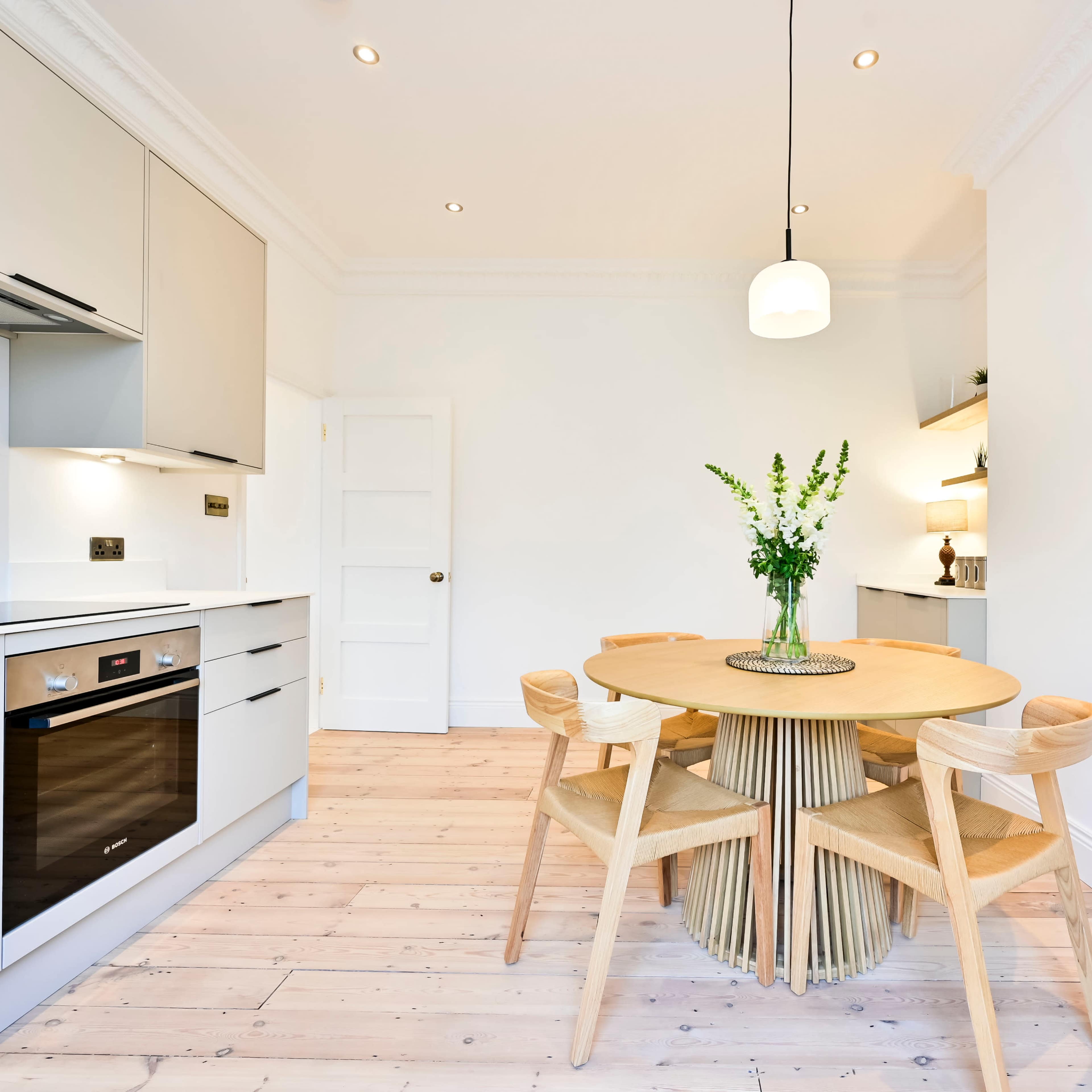 A modern kitchen features a circular wooden dining table surrounded by four chairs, with a vase of flowers in the center.