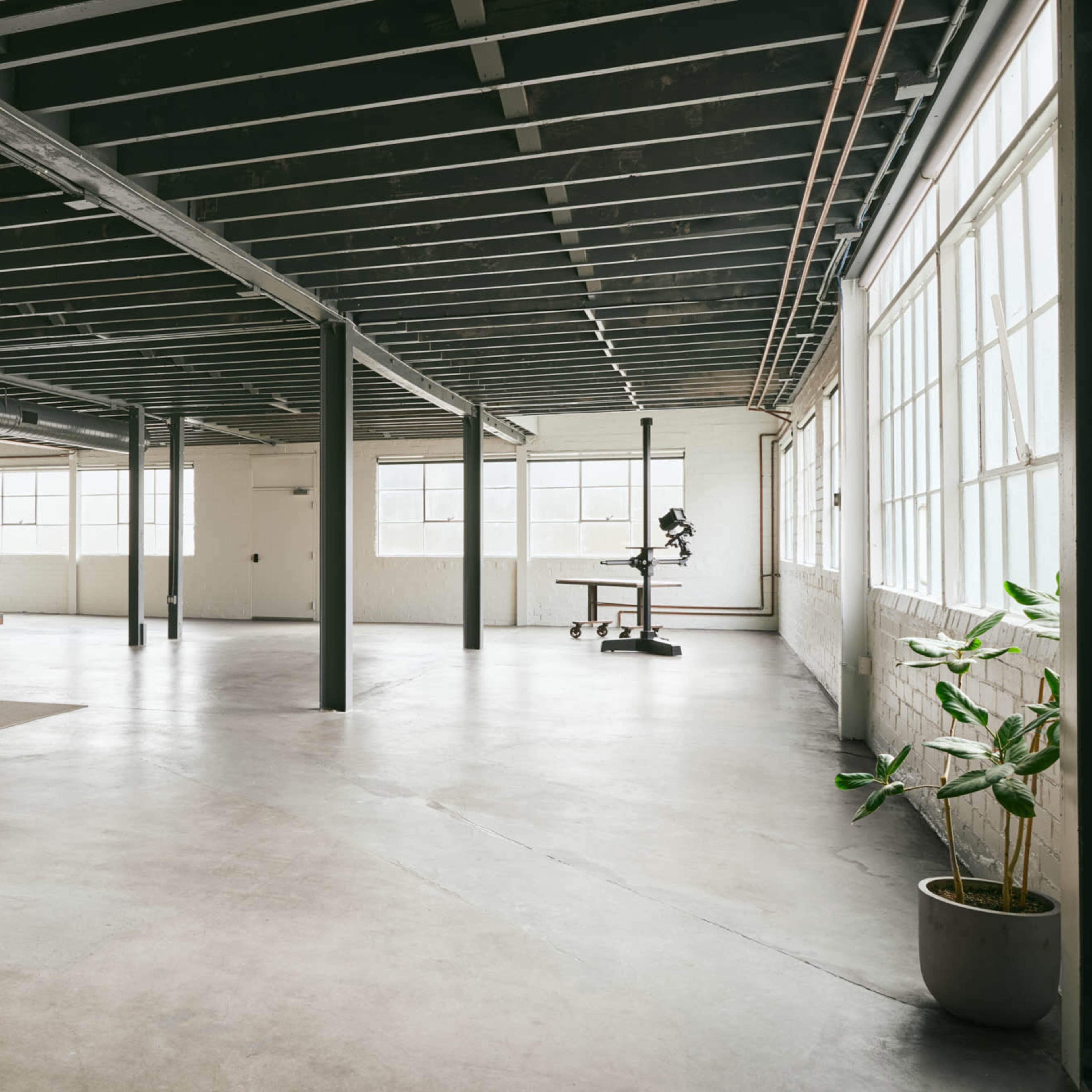 A spacious, industrial-style office with large windows, concrete floors, a meeting table with chairs, and a corner desk with an ergonomic chair.