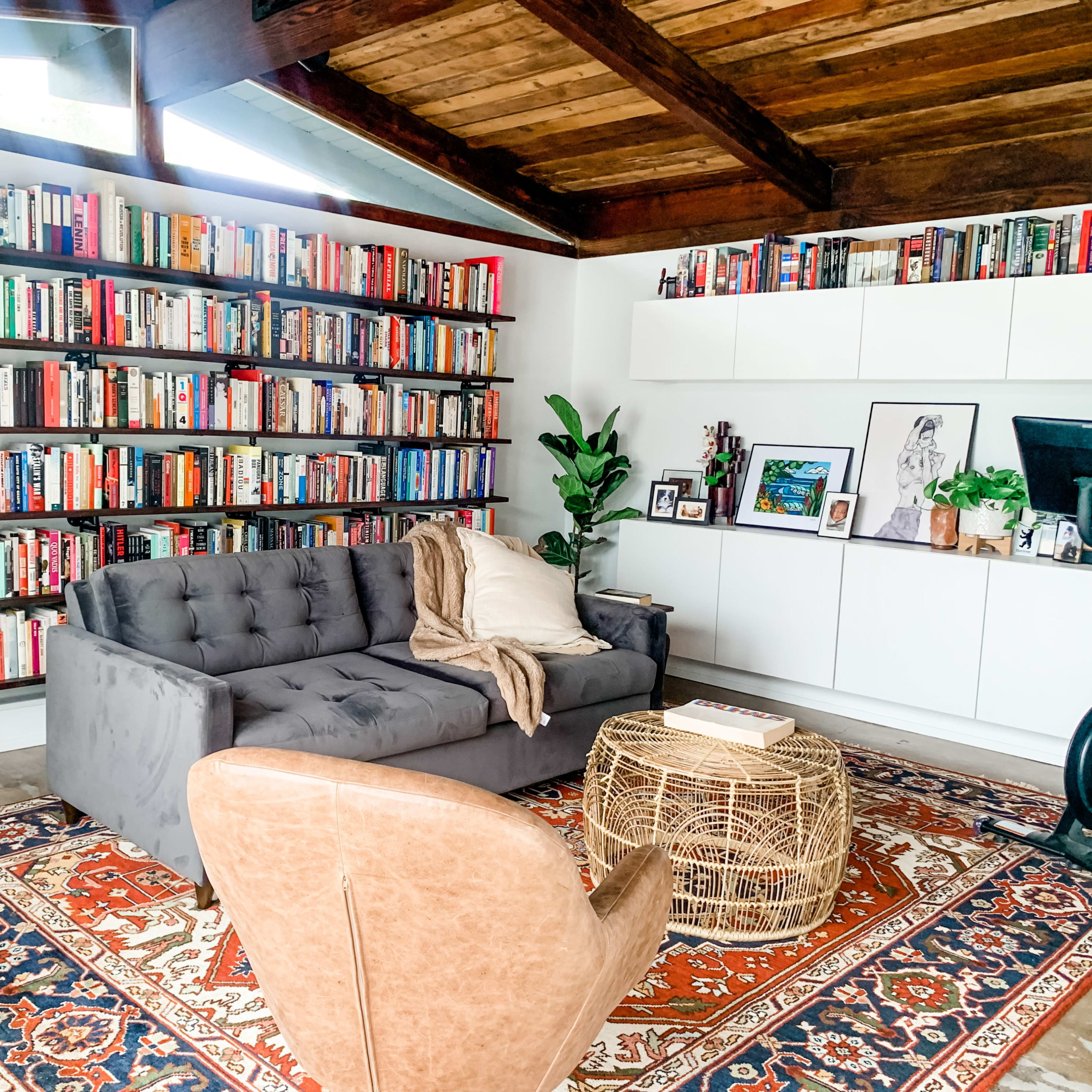 A cozy living room features a large bookshelf filled with books, a gray sofa, a round wicker coffee table, and a single leather chair on a patterned rug.