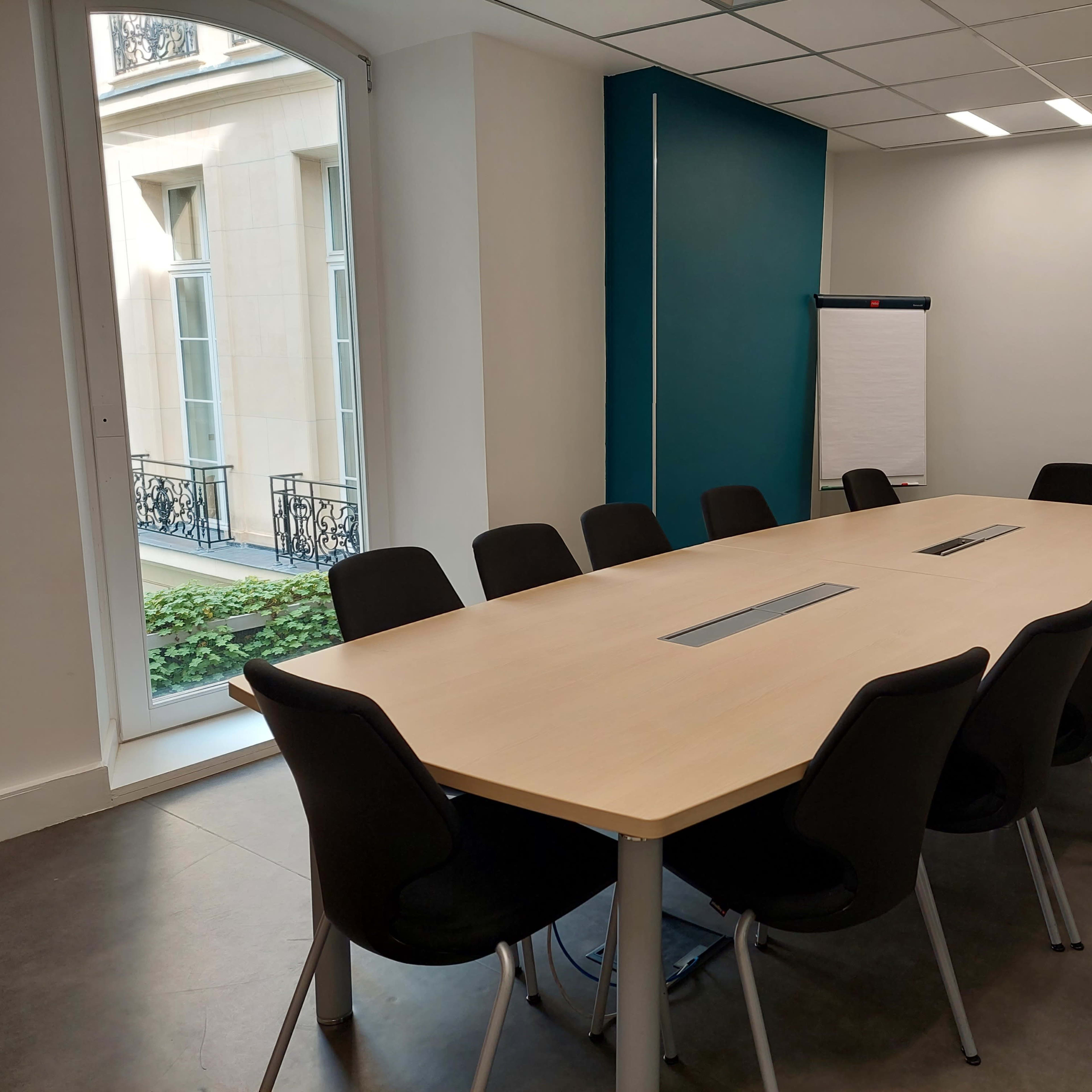 A modern conference room with a long wooden table surrounded by black chairs, featuring a flip chart and large windows letting in natural light.