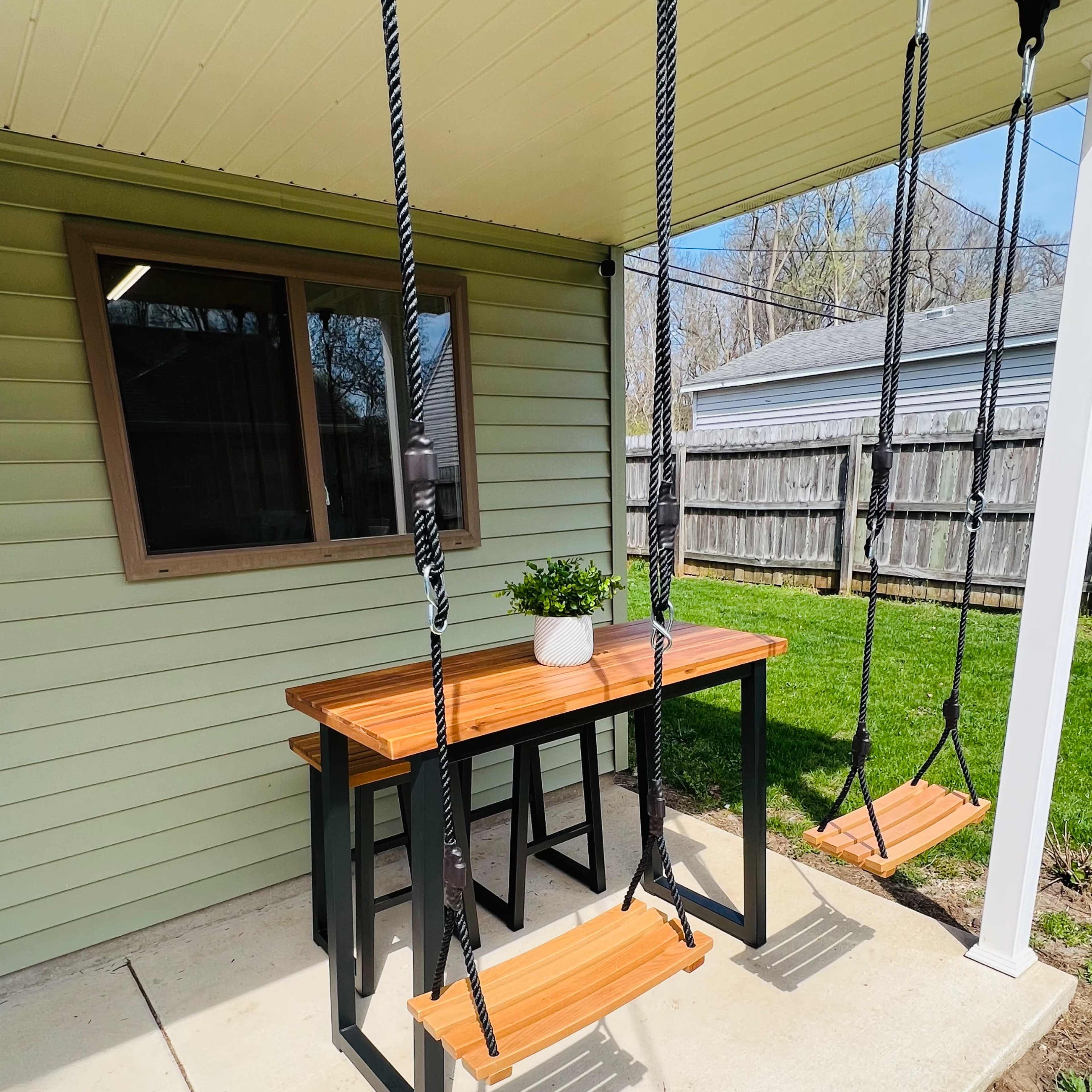 A table with two swings attached is set under a covered patio next to a grassy area.