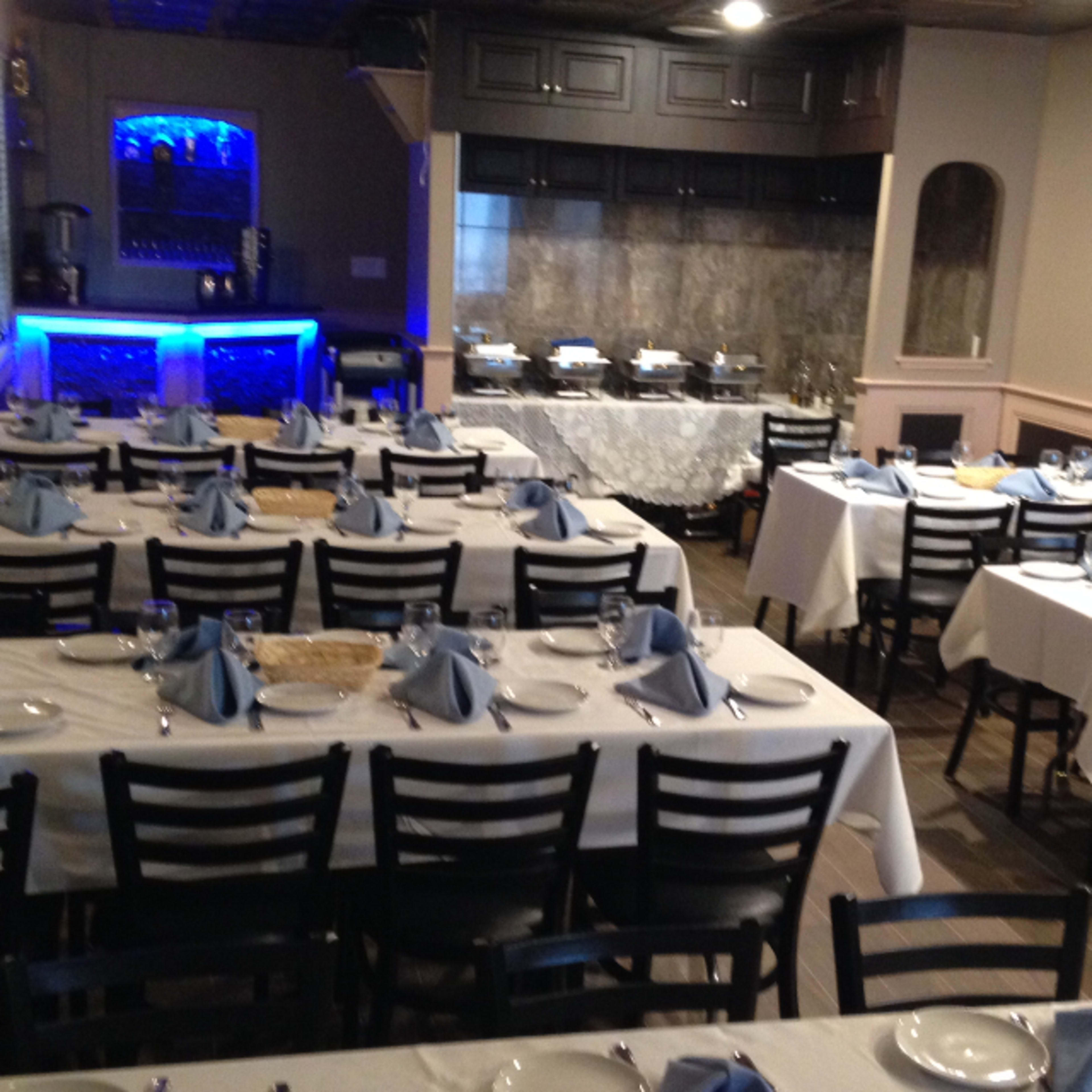 A dining room set with multiple tables dressed in white tablecloths and blue napkins, with a bar area in the background.