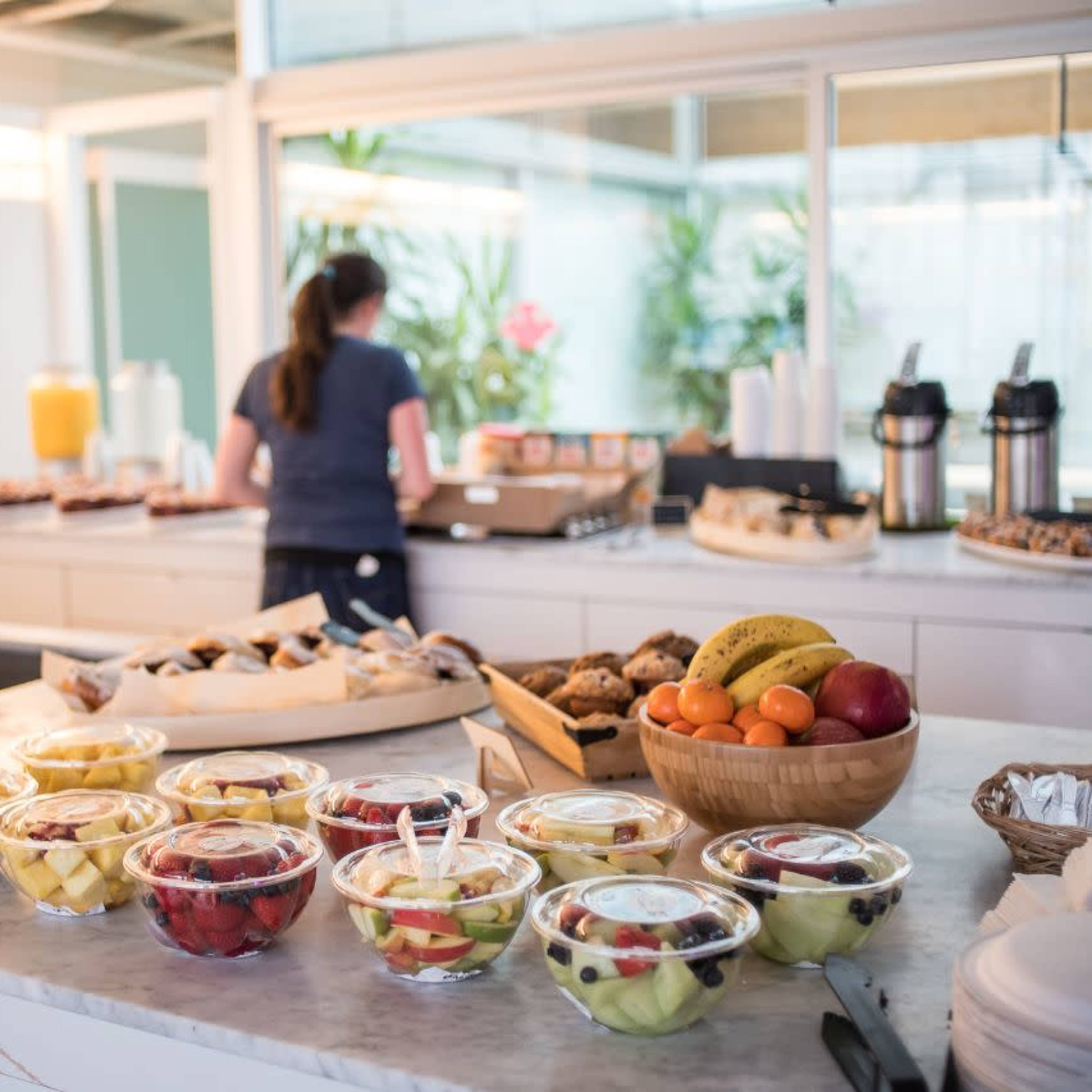 A buffet table displays an assortment of fruit cups and baked goods, while a person prepares food in the background.