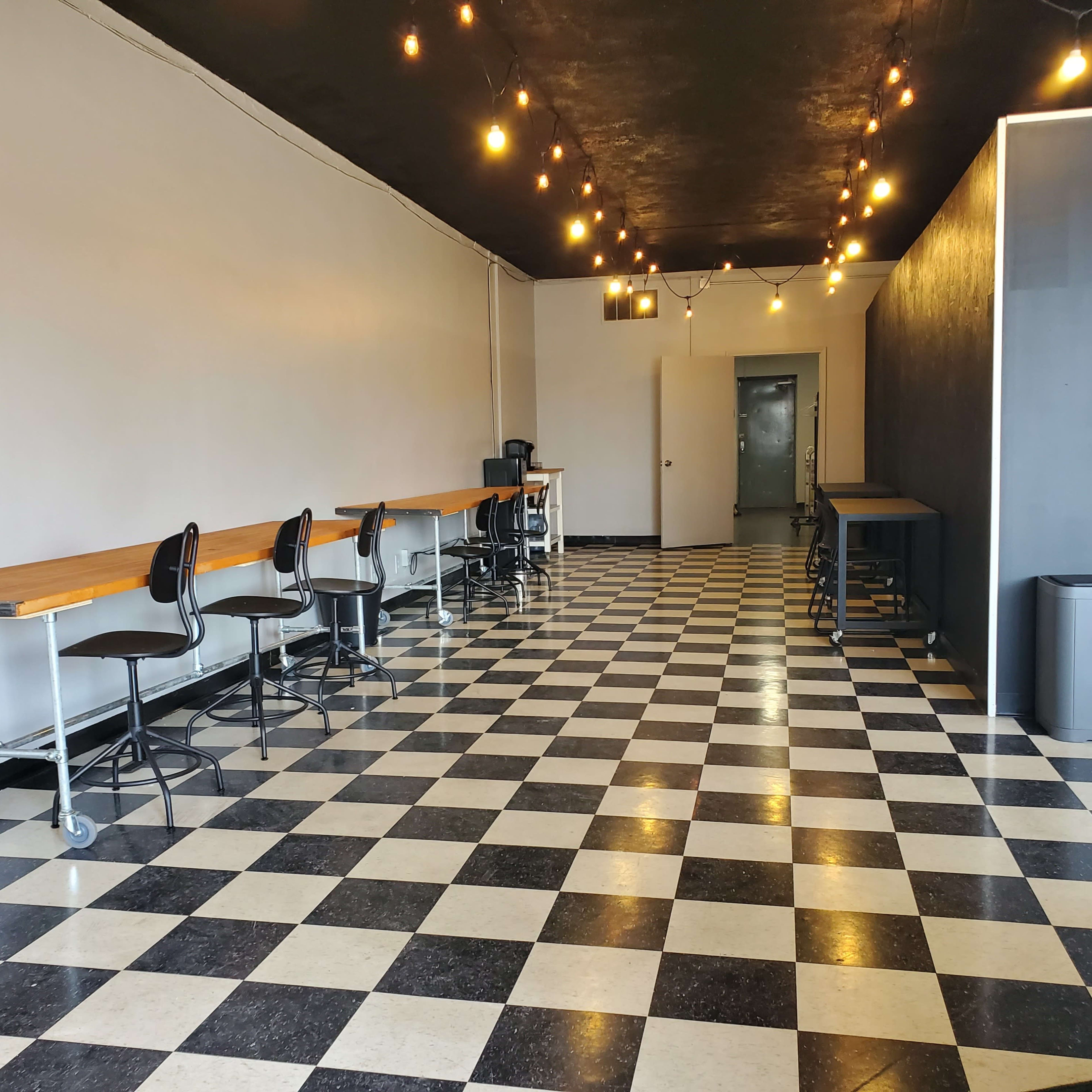 A spacious interior with black and white checkered flooring, featuring several bar-height stools at long tables and pendant lights hanging from the ceiling.