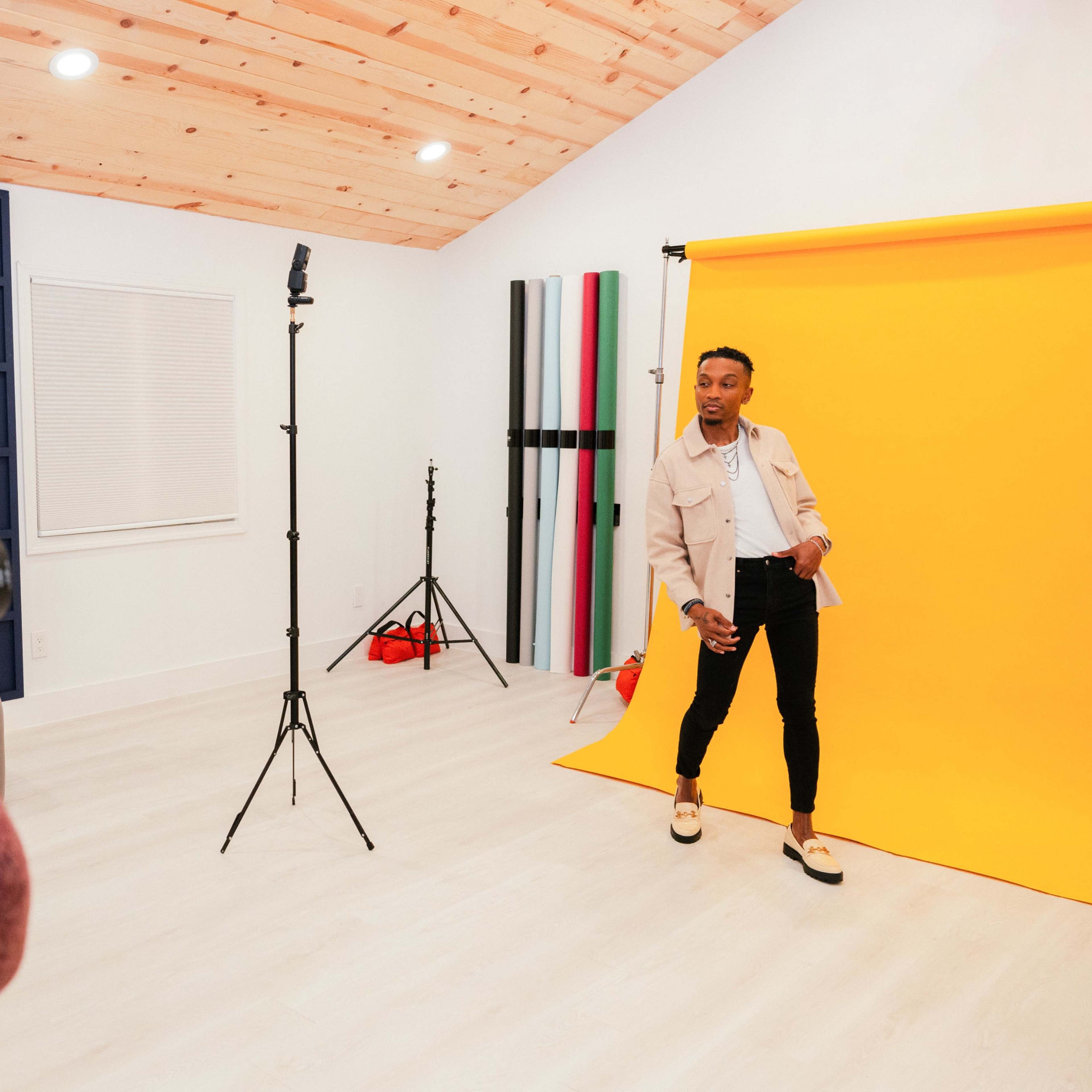 A person poses in front of a bright yellow backdrop while a photographer takes their picture in a well-lit studio.