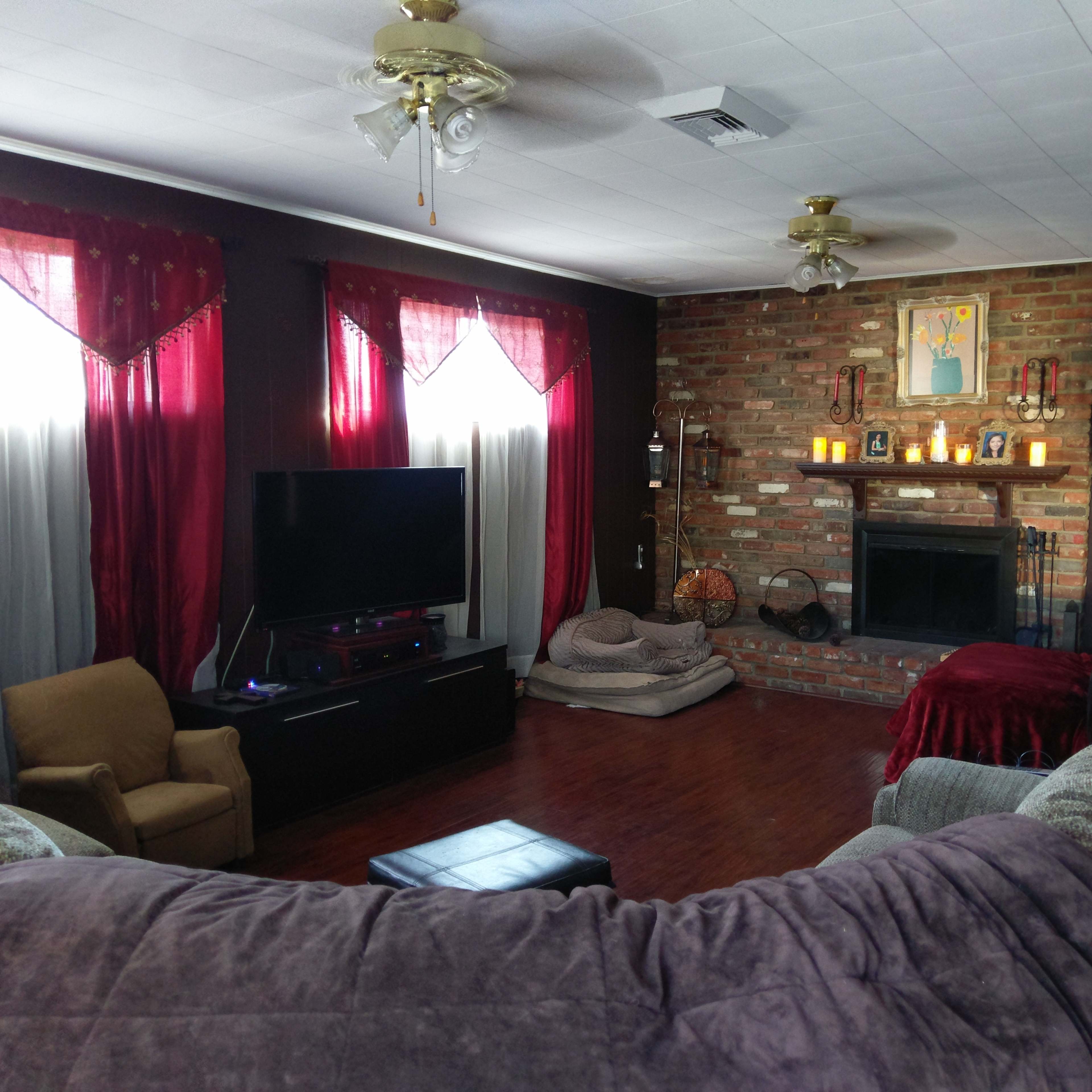 The living room features a sectional sofa, a television unit, a fireplace with candles, and large windows adorned with red curtains.