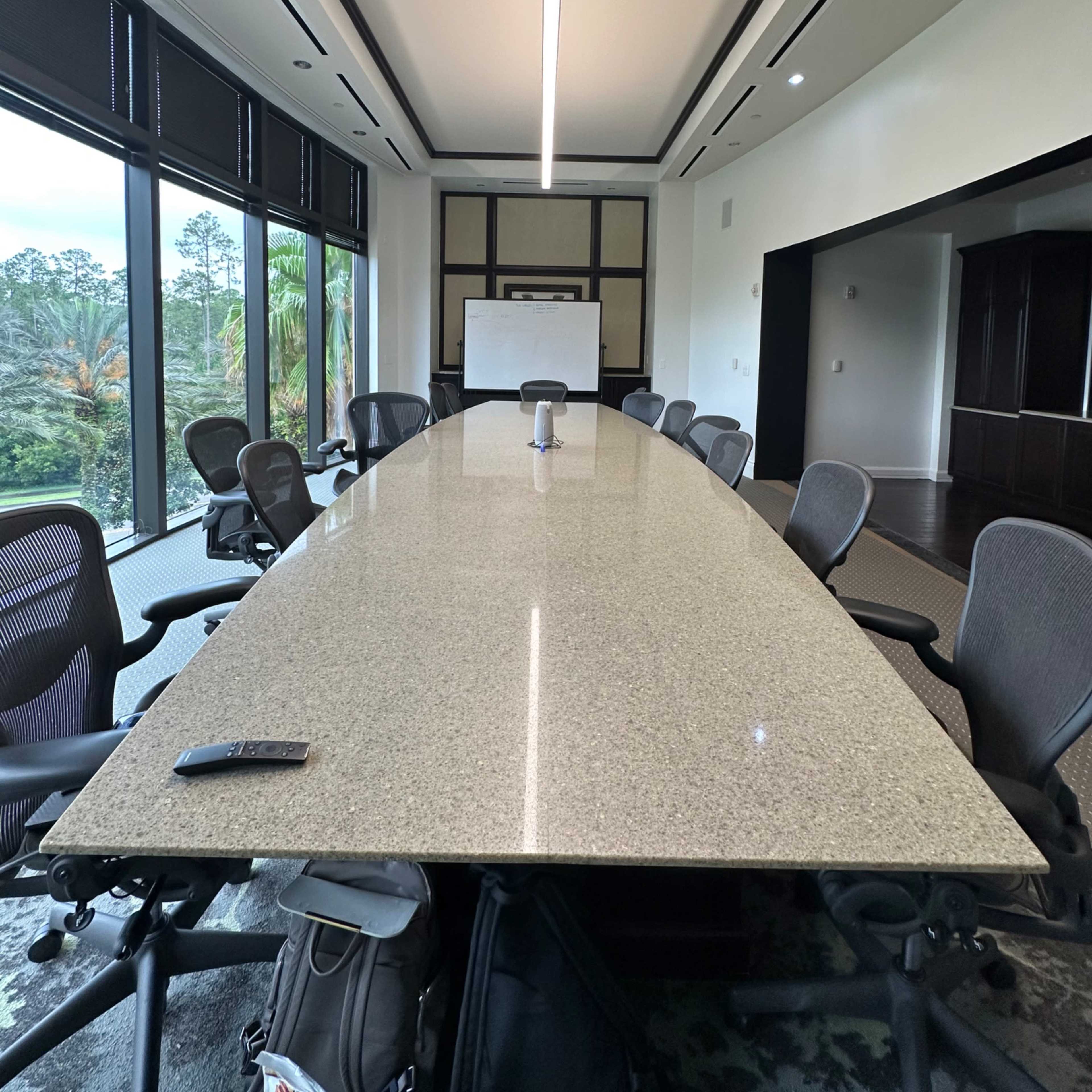 A large, modern conference room with a long, smooth table surrounded by office chairs and large windows overlooking a green landscape.