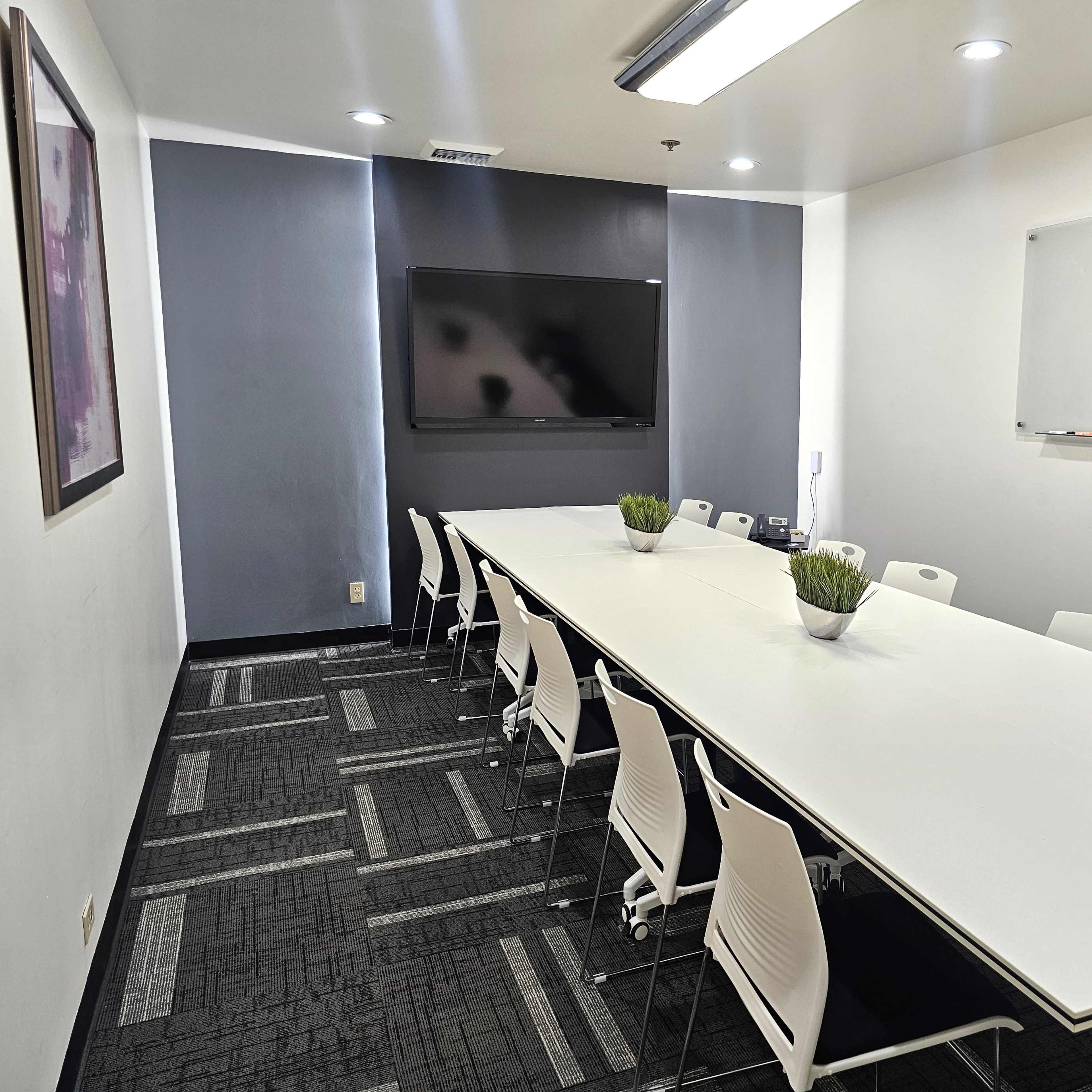 A long conference table with white chairs is set up in a modern meeting room featuring a television and a whiteboard.