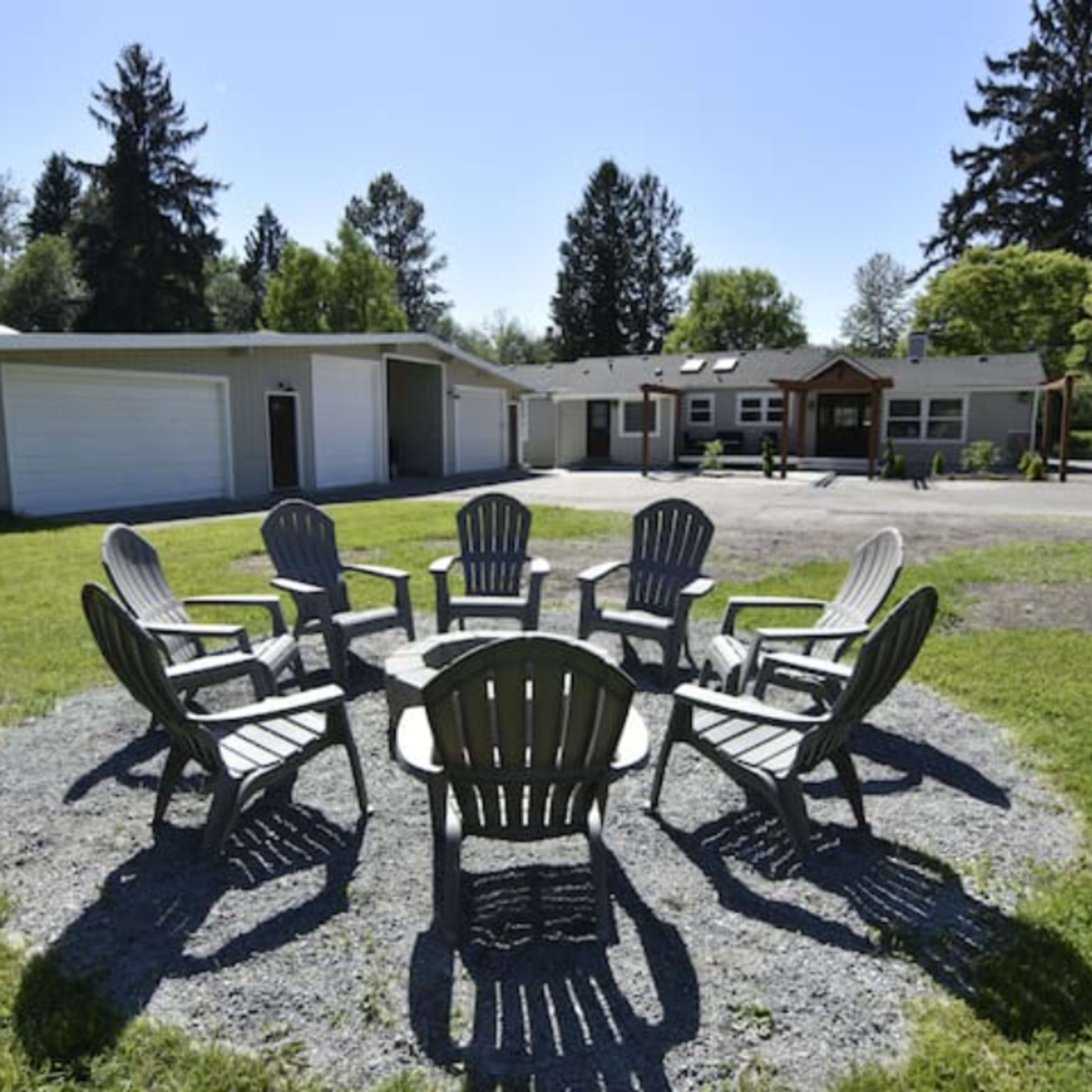 A circular arrangement of eight Adirondack chairs surrounding a stone fire pit in front of a single-story house and garage, set in a grassy area with trees in the background.