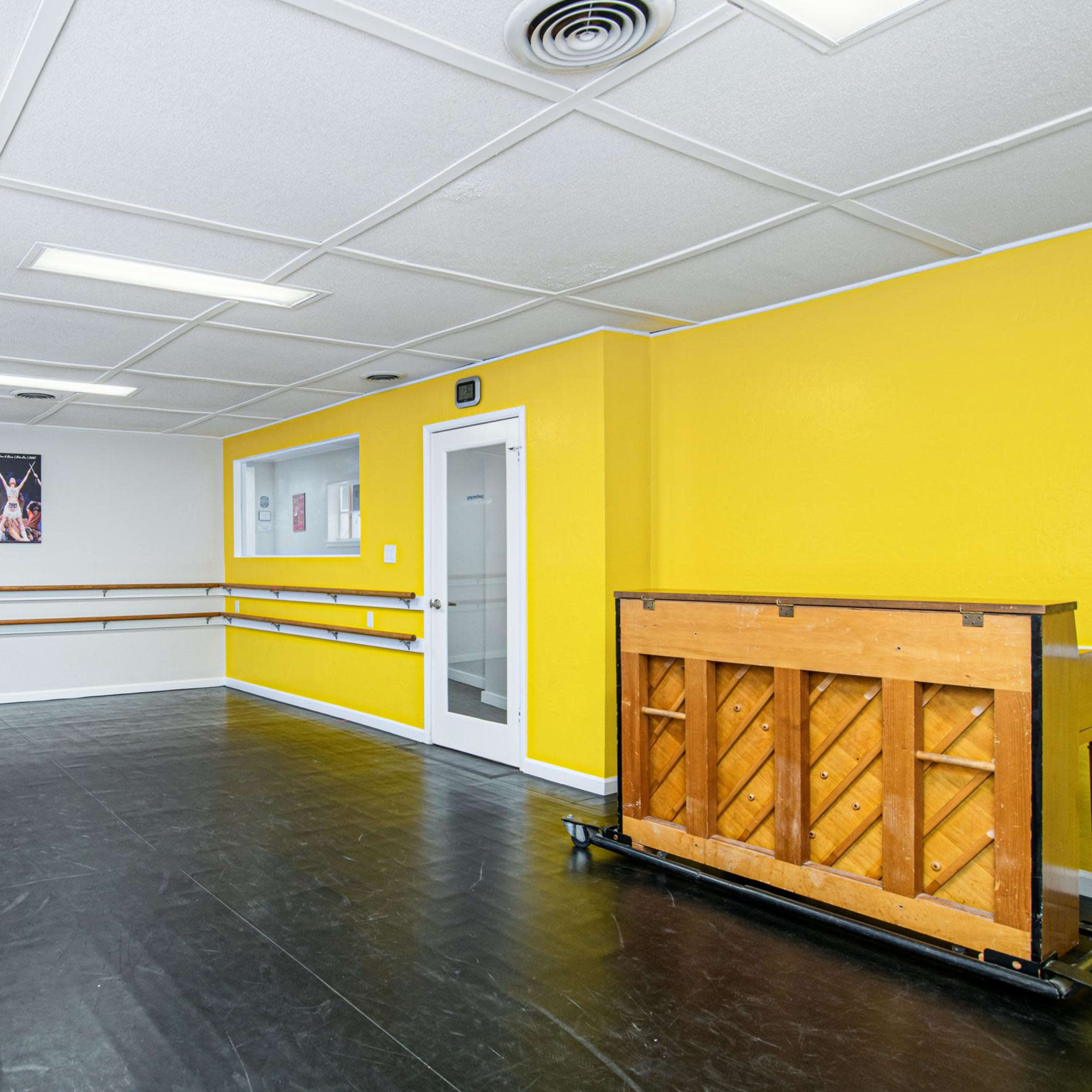 A spacious dance studio featuring a yellow wall, ballet barres along one side, and a wooden piano against another wall.
