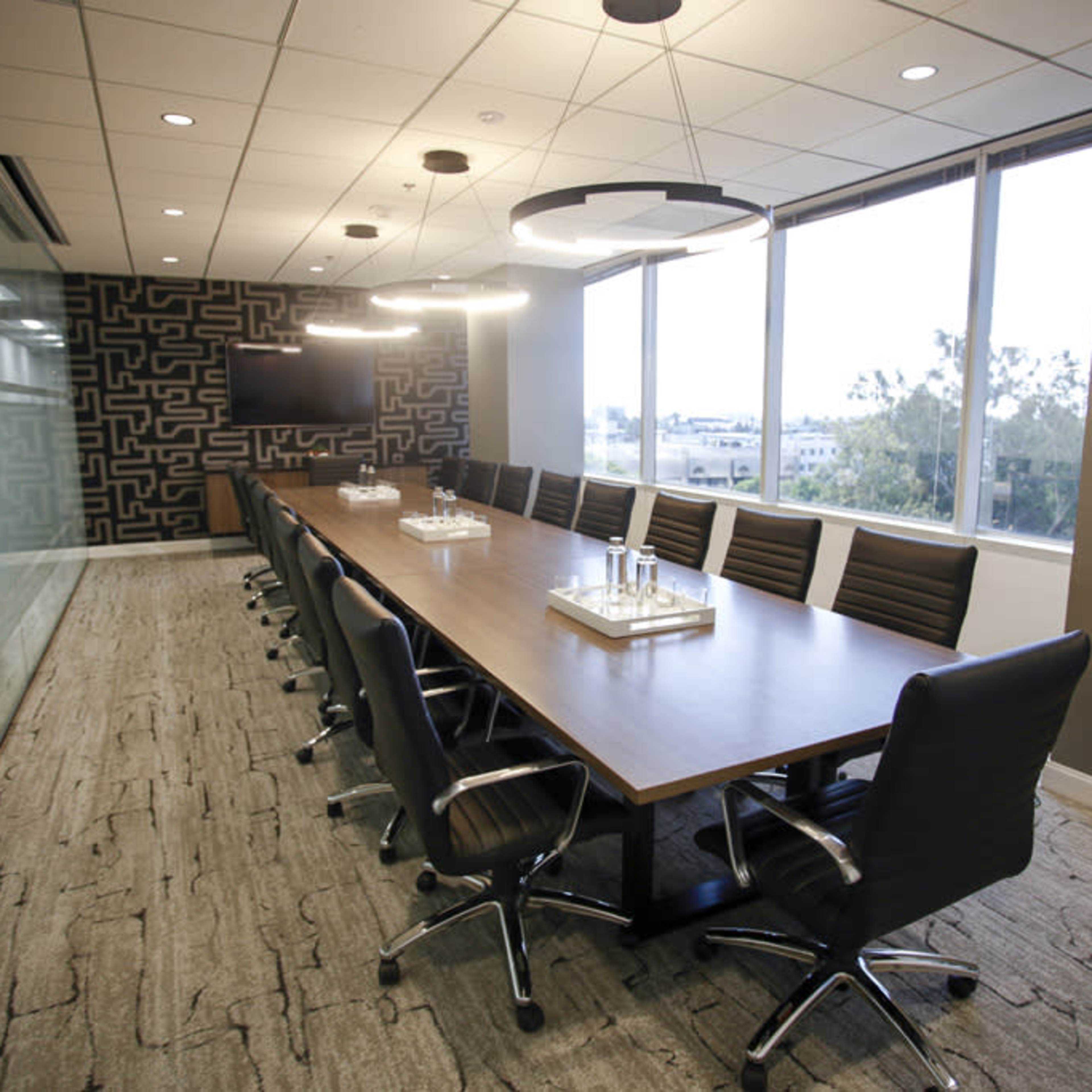 A long conference table with ergonomic chairs is positioned in a modern meeting room featuring large windows and a patterned accent wall.