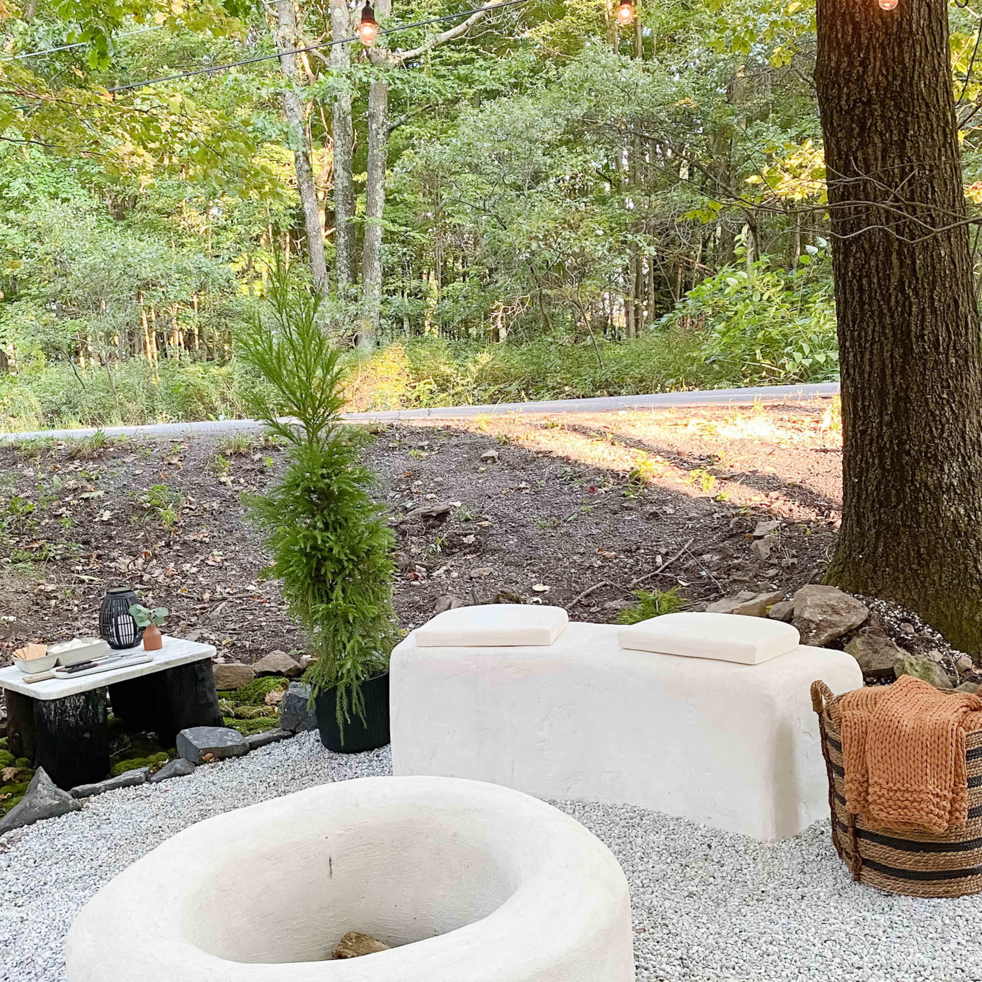 The scene features a minimalist outdoor seating area with a circular fire pit, two solid benches, and a small table surrounded by gravel and trees.