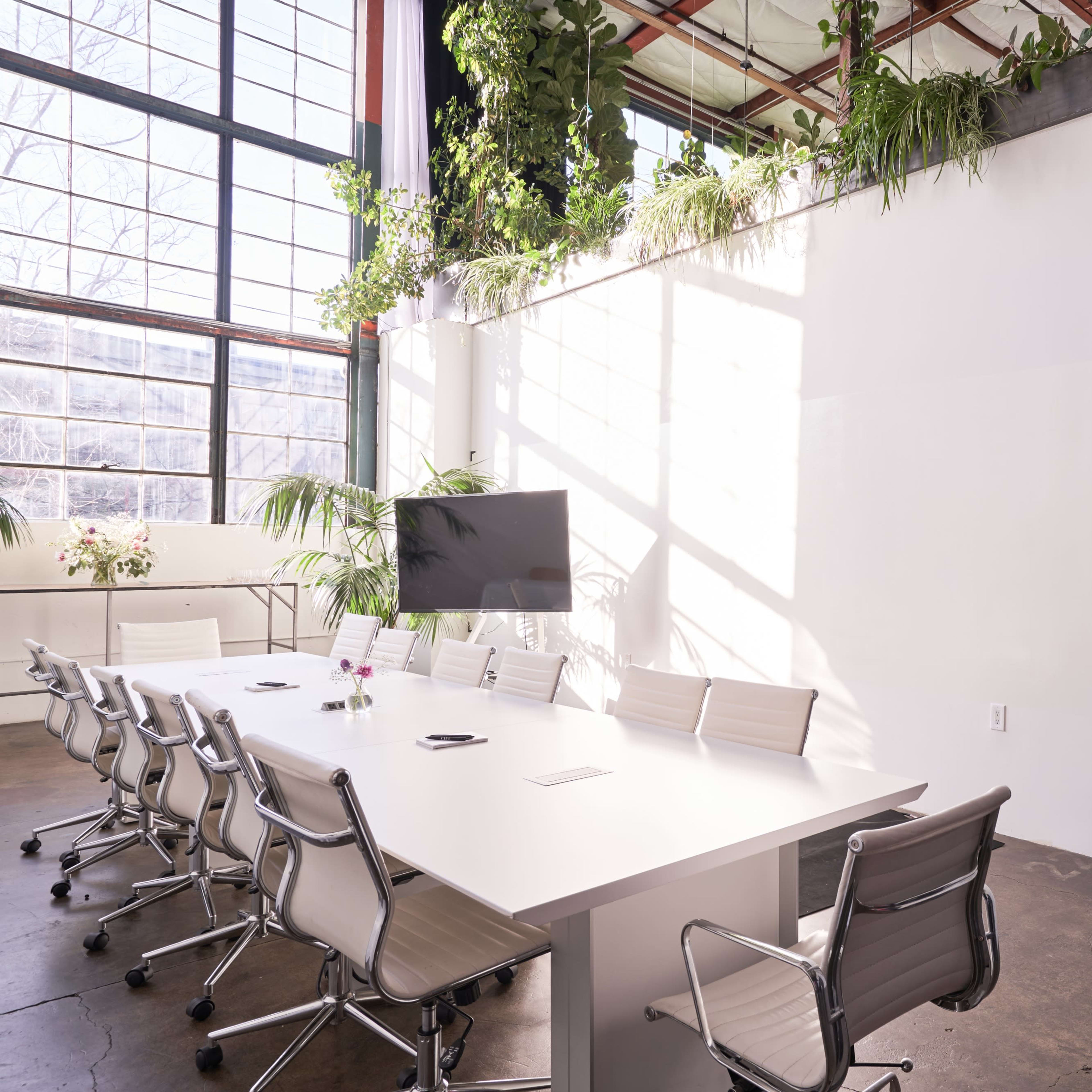 A bright conference room features a long white table surrounded by modern chairs, with large windows and plants lining the walls.