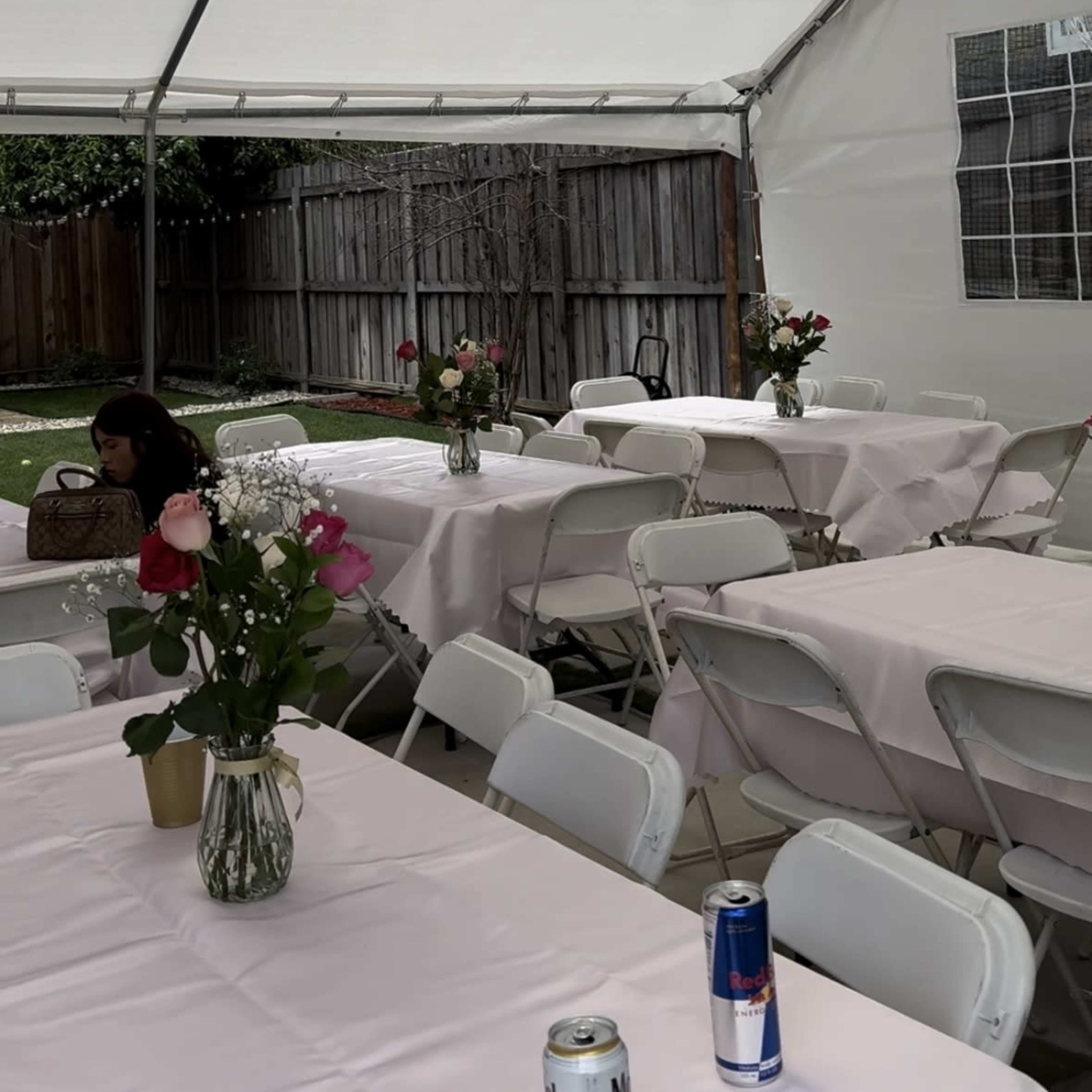 A large white tent is set up in a backyard, with several tables covered in pink tablecloths, each adorned with flower arrangements, while a person sits alone at one of the tables.