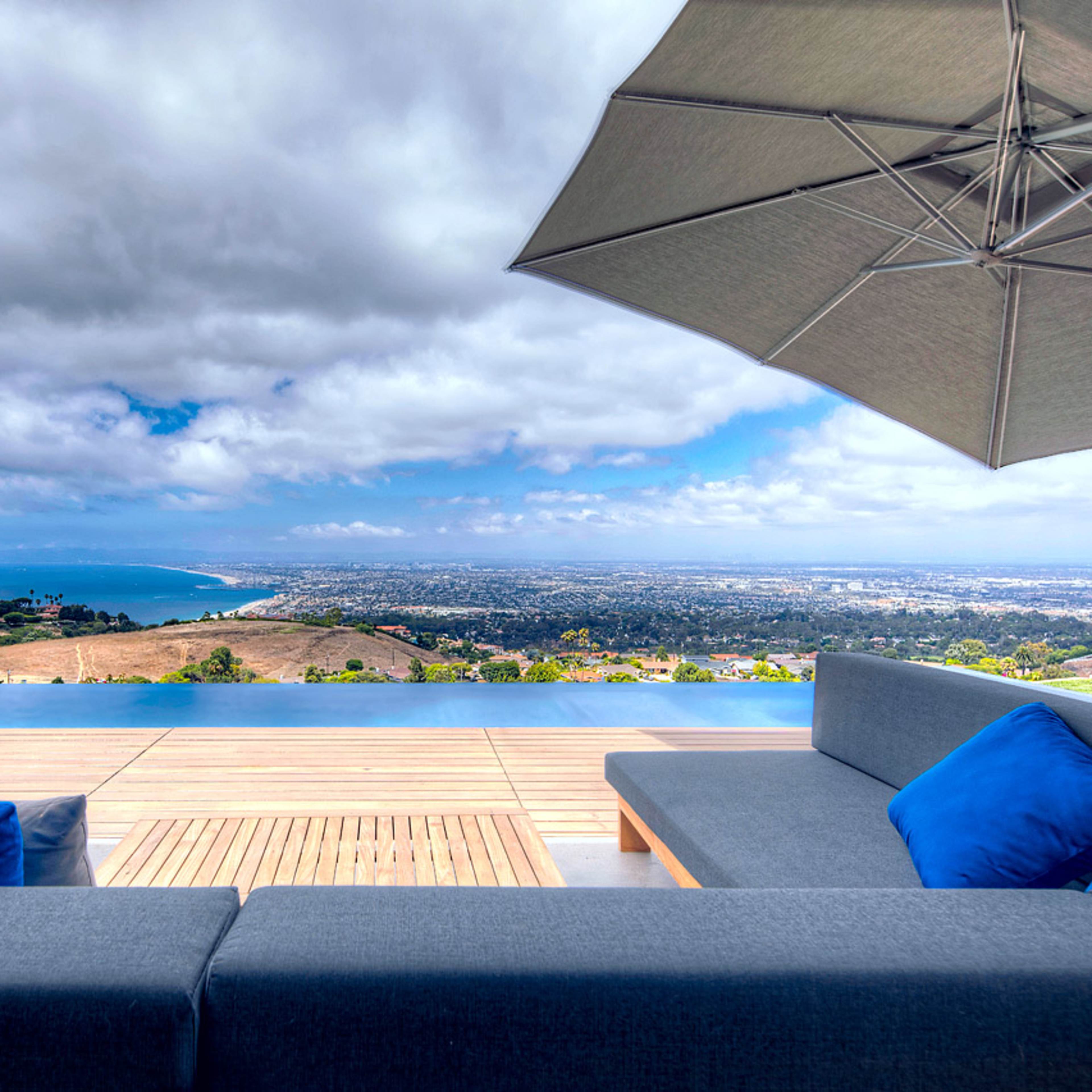 A modern outdoor lounge area features a sectional sofa with blue cushions, overlooking a panoramic view of the ocean and landscape under a cloudy sky.