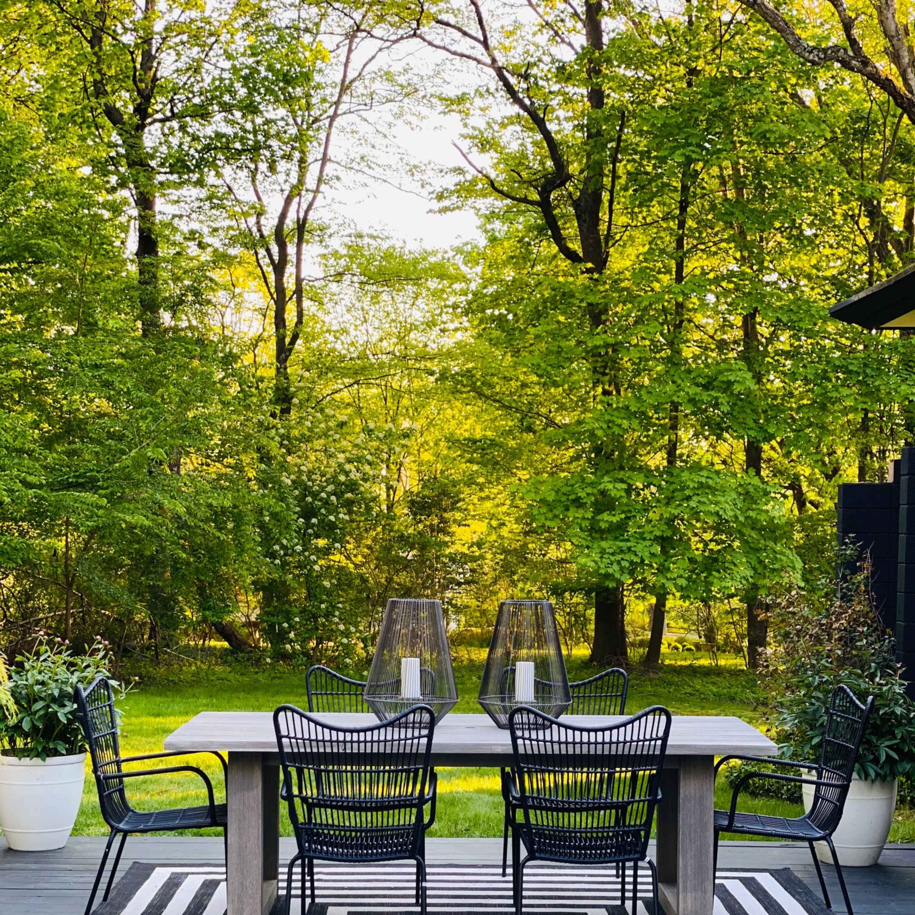 A wooden dining table is set on a black and white striped rug, surrounded by black chairs, with greenery and trees in the background.