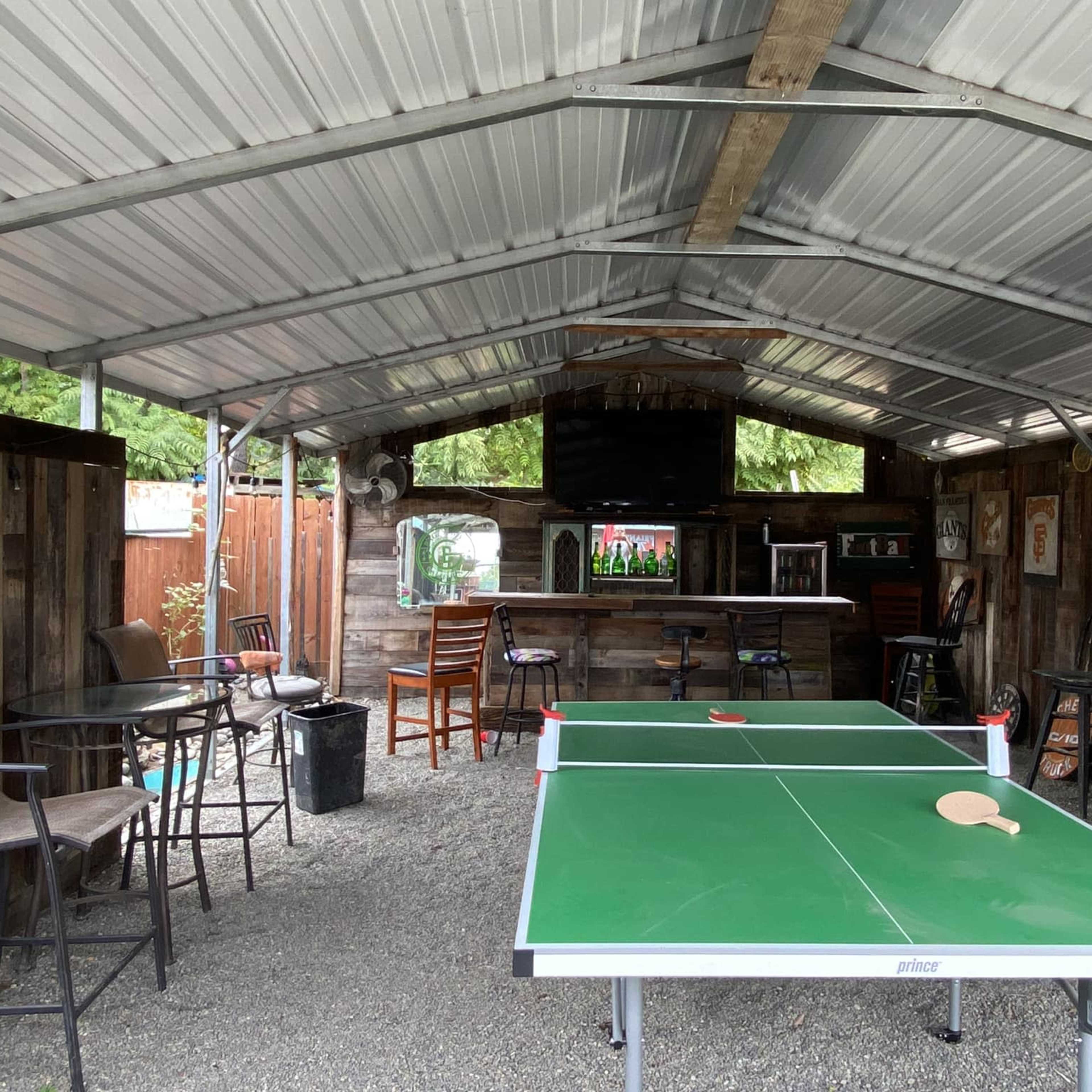 A covered outdoor area with a green ping pong table and bar seating against wooden walls and a television in the background.