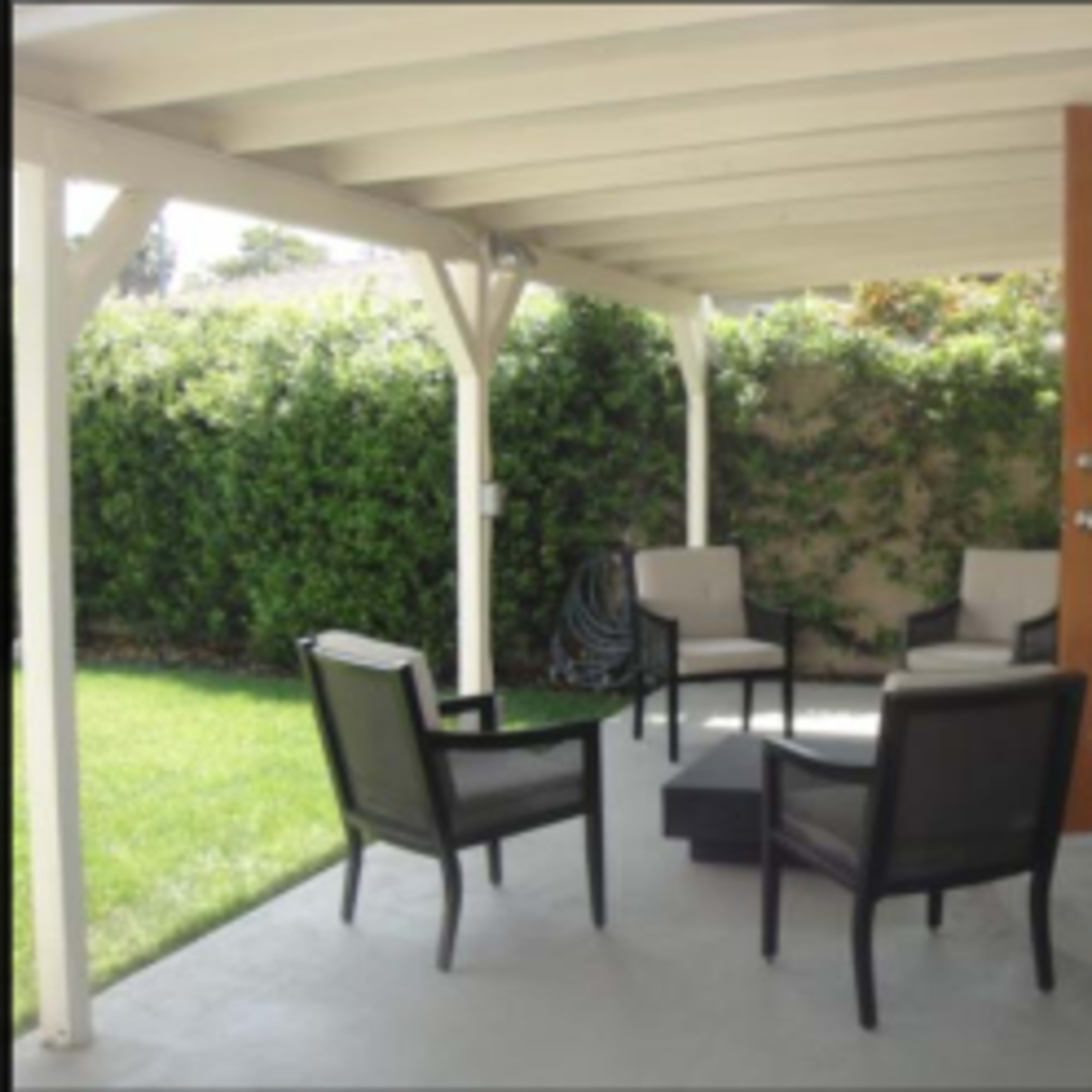 A covered patio features four chairs arranged around a low table, with a grassy area and hedge in the background.