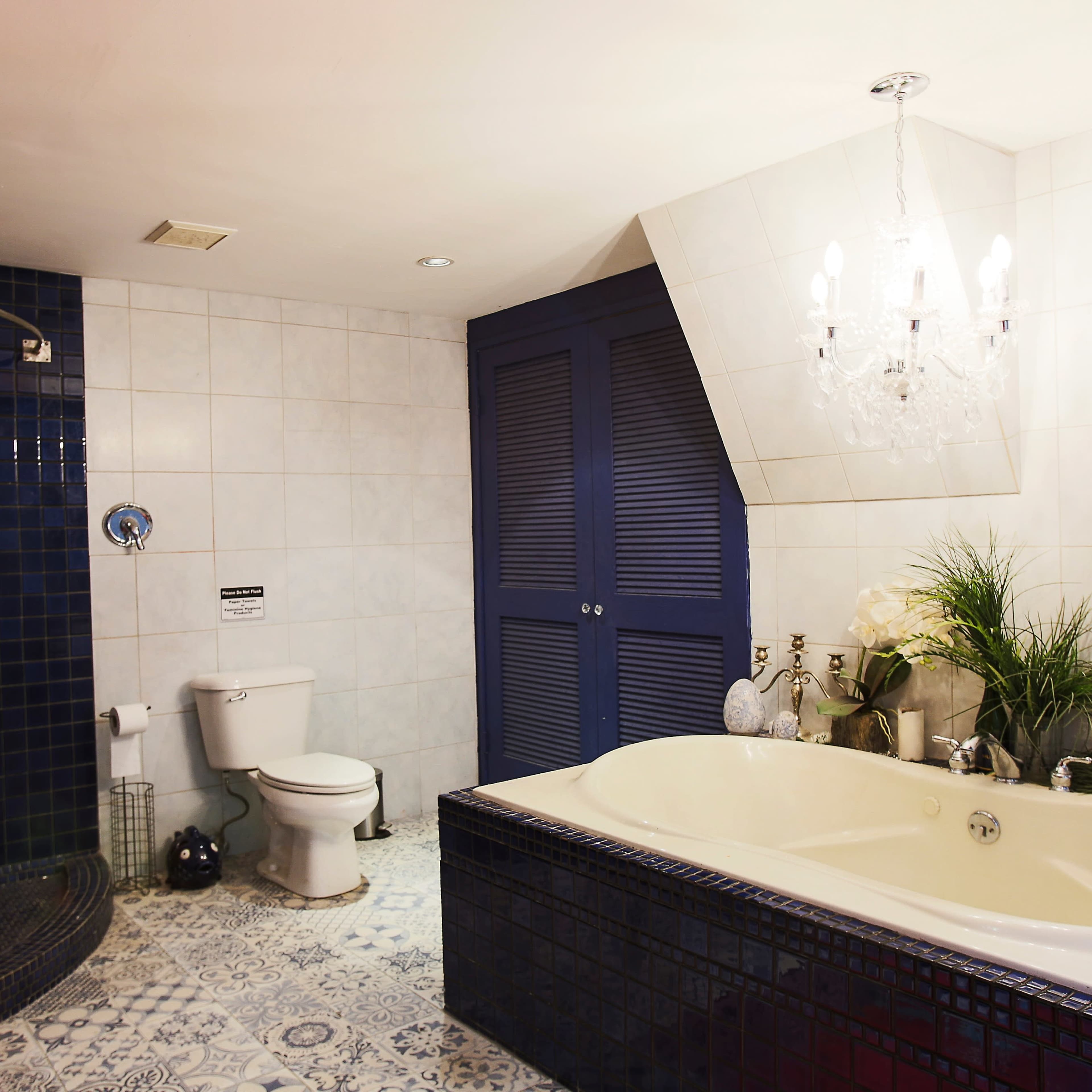 A bathroom featuring a shower area, a white toilet, a double sink with a blue tiled wall, and a large bathtub, all set against light-colored tiles and illuminated by a chandelier.