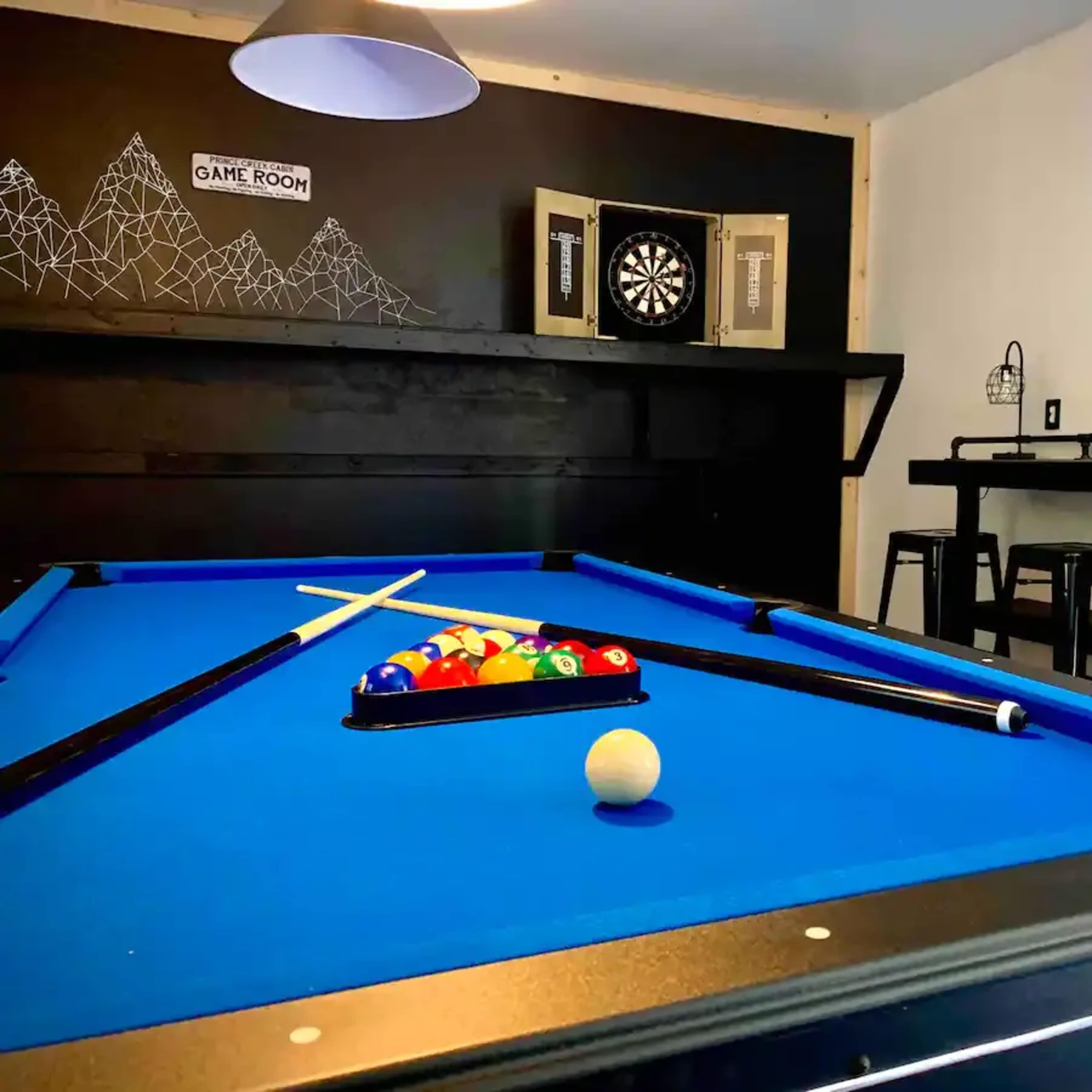 A game room featuring a blue pool table with balls arranged in a rack, a dartboard on the wall, and a bar-height table with stools.
