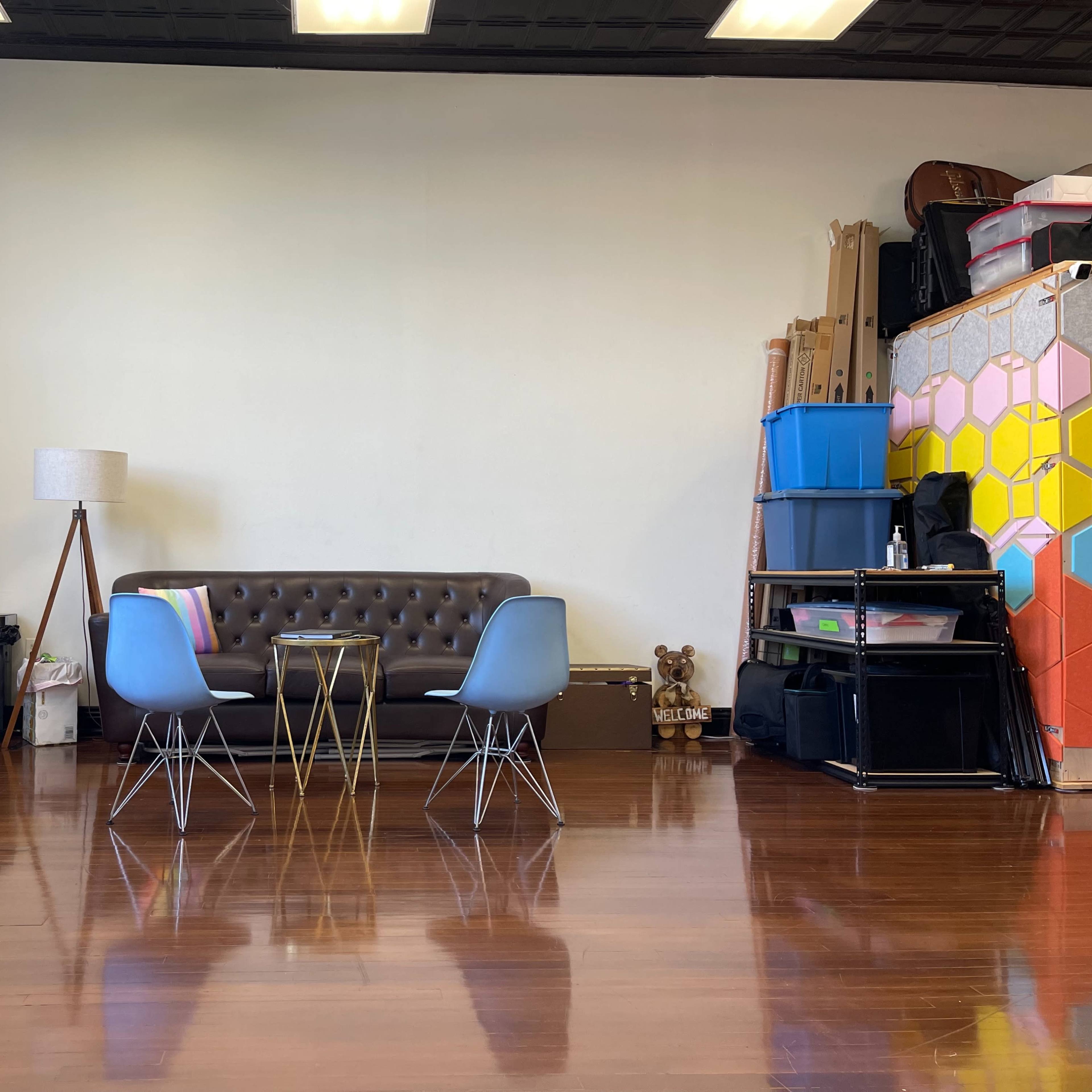 A spacious room with hardwood floors, featuring a brown sofa, two chairs, a small coffee table, and various storage items along the walls.