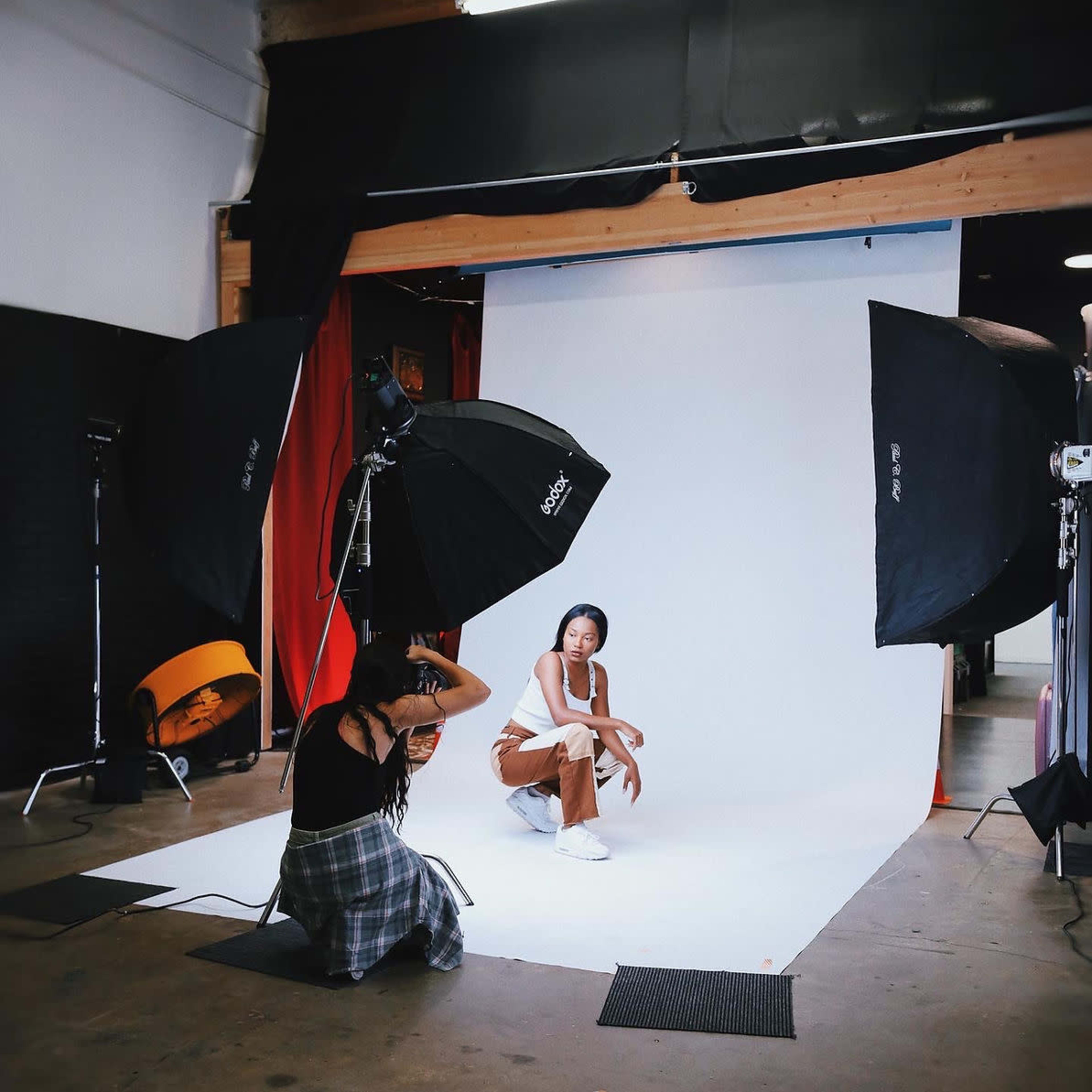 A model poses on a white backdrop while a photographer captures the shot, surrounded by studio lighting equipment.
