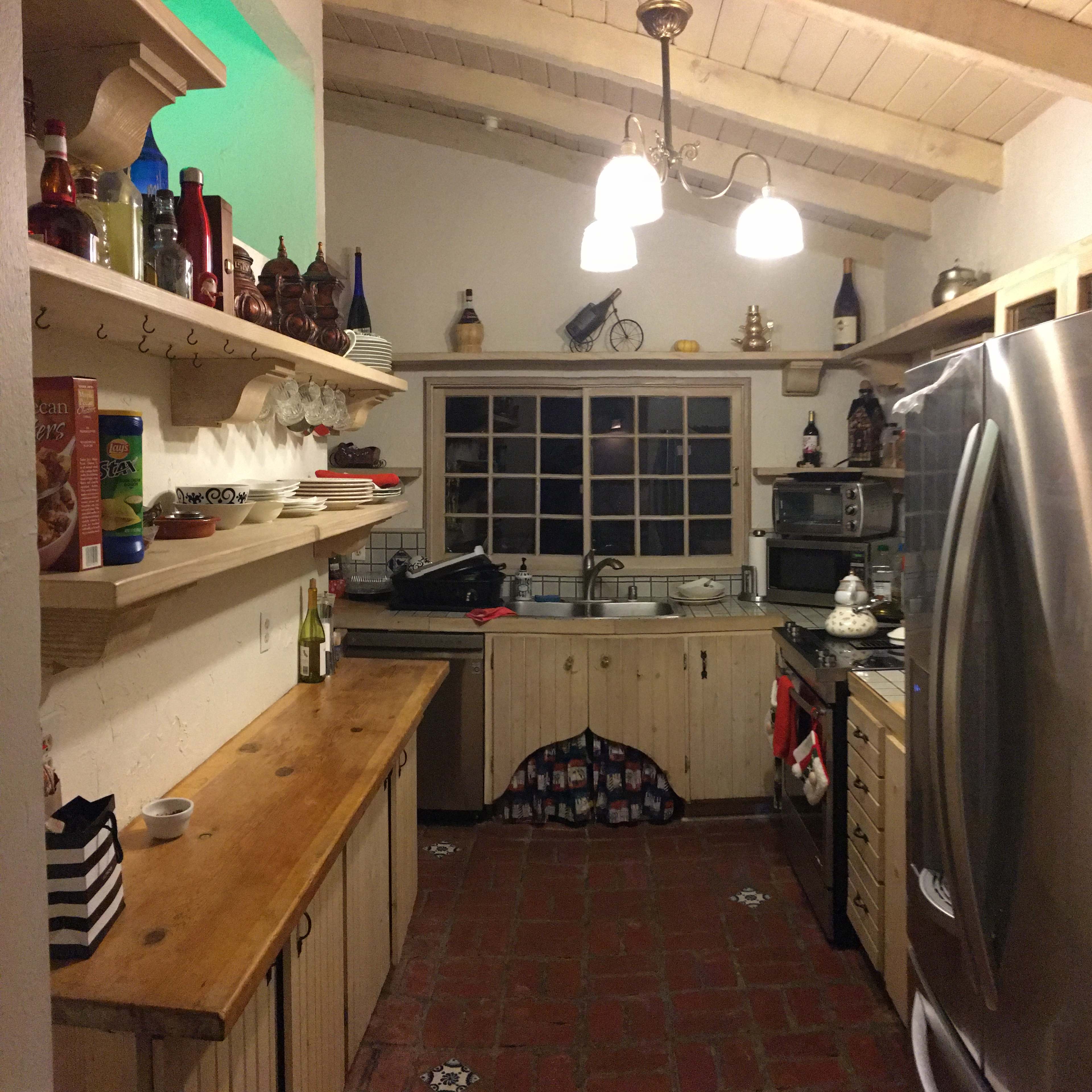 A small kitchen features wooden shelves stocked with various bottles and dishes, a stainless steel refrigerator, and a window above the sink.