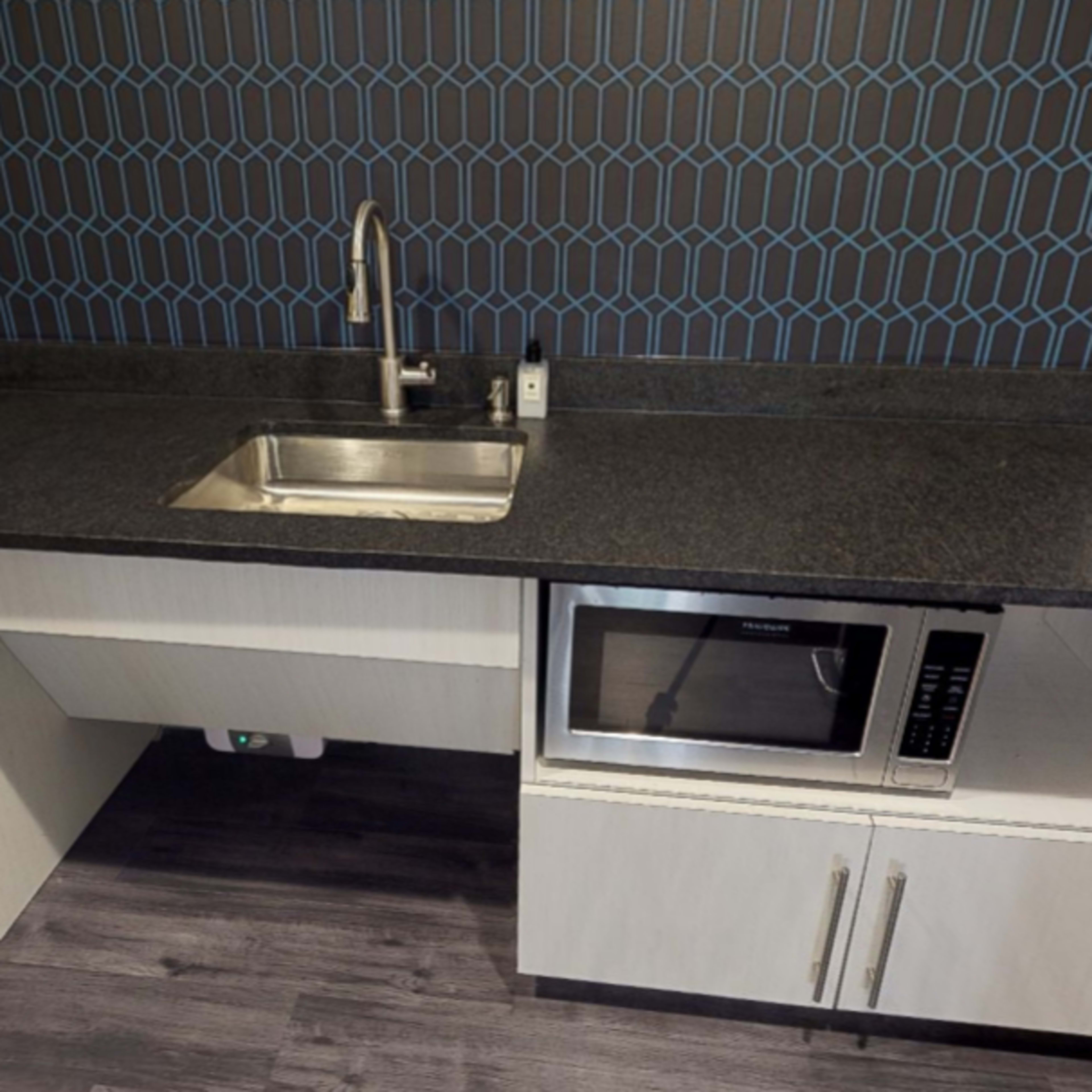 A modern kitchen area featuring a sink, microwave, and a small refrigerator beneath a dark countertop with a patterned backsplash.