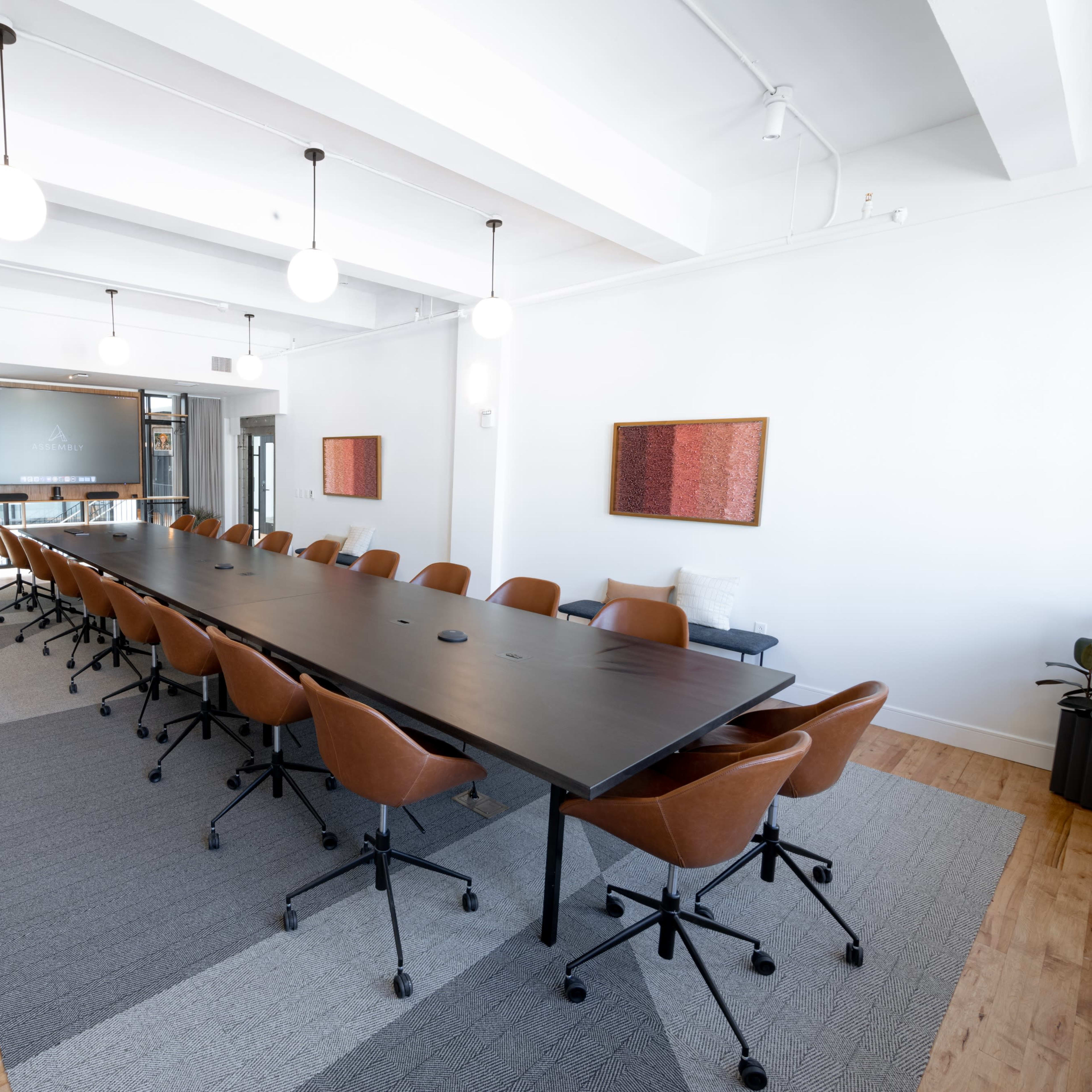 A long conference table with brown chairs is set up in a well-lit meeting room featuring large windows and modern decor.
