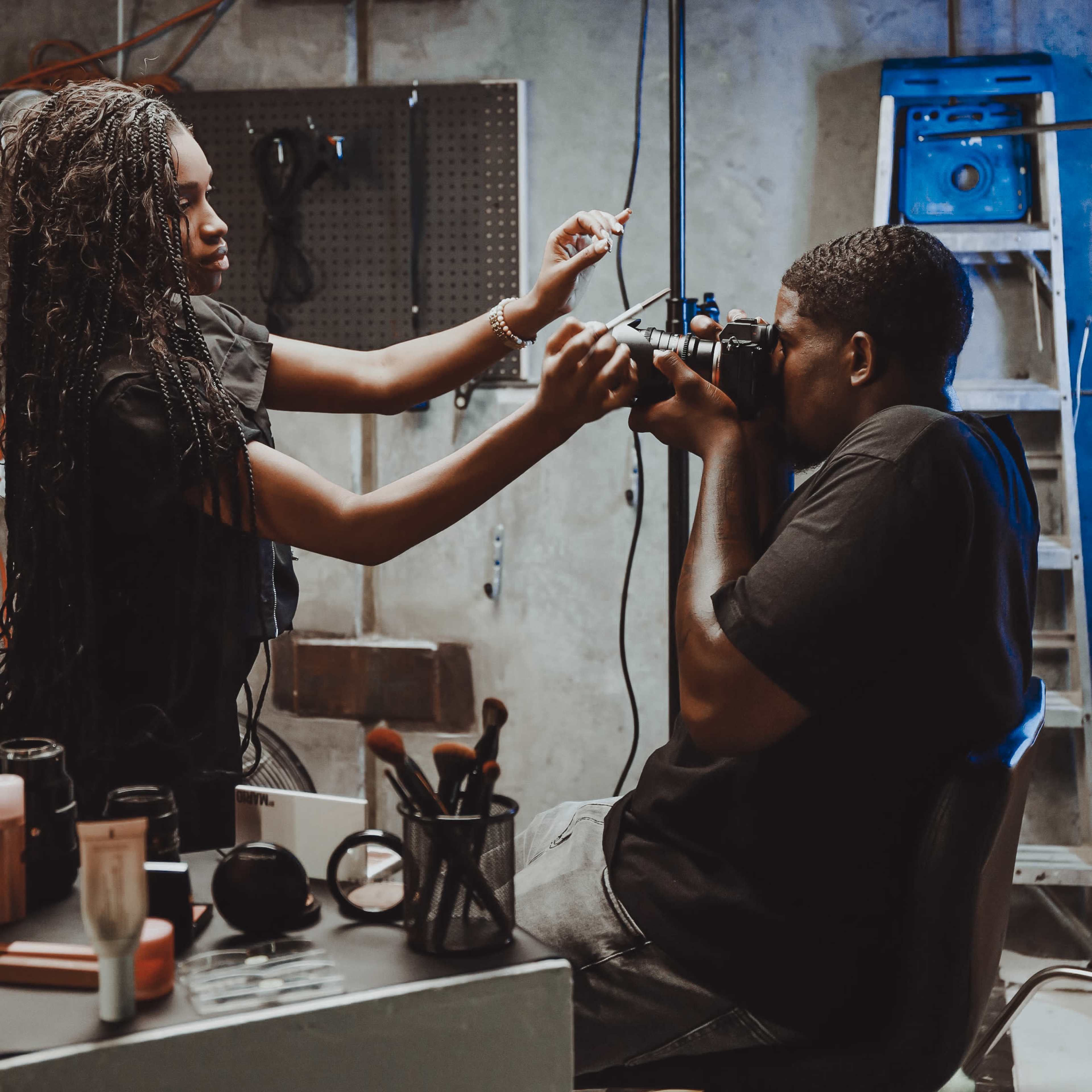 A woman adjusts the camera while a man sits in a chair, preparing to take a photograph in a studio setting.