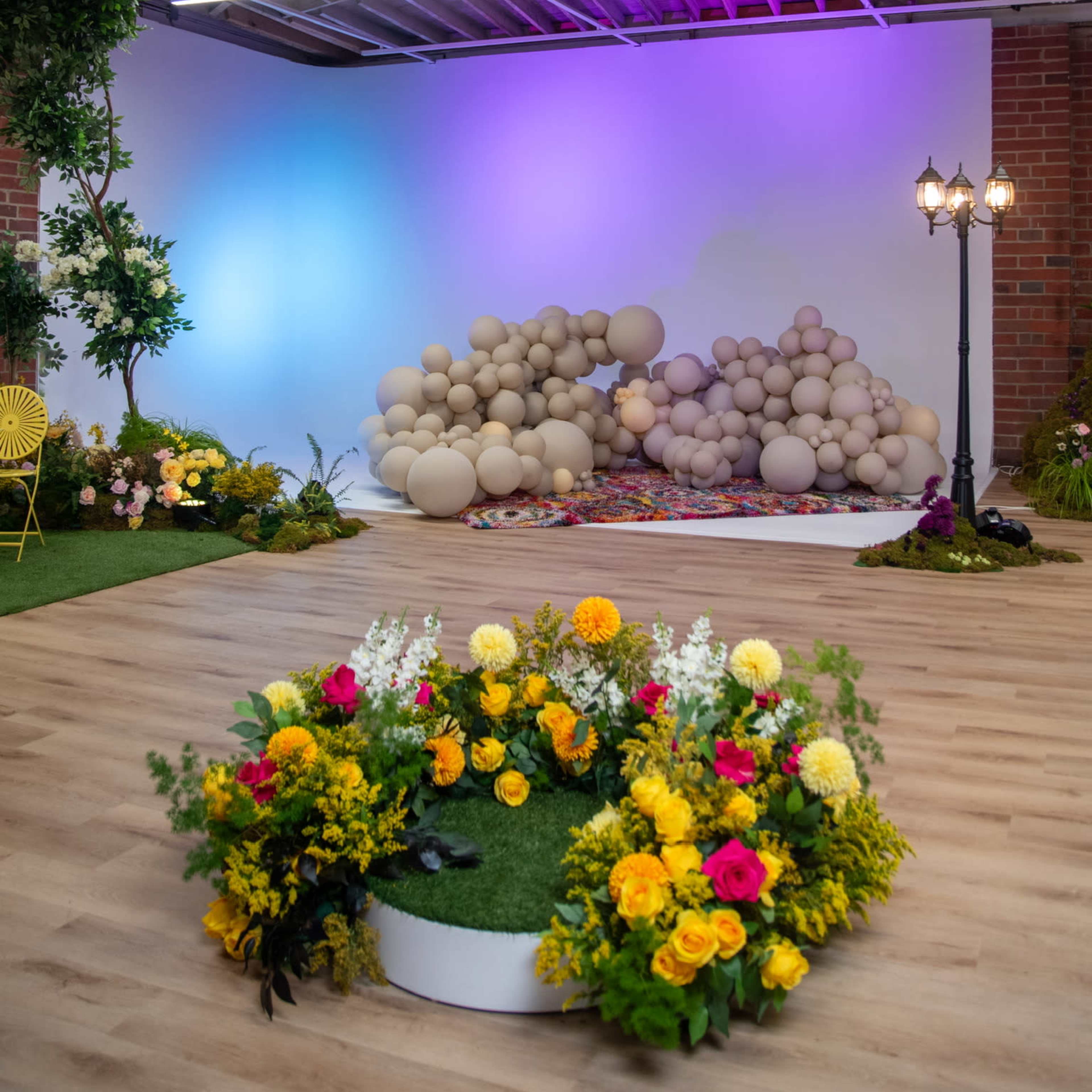 The scene features a decorated studio space with colorful floral arrangements, a large balloon installation, and vibrant lighting.