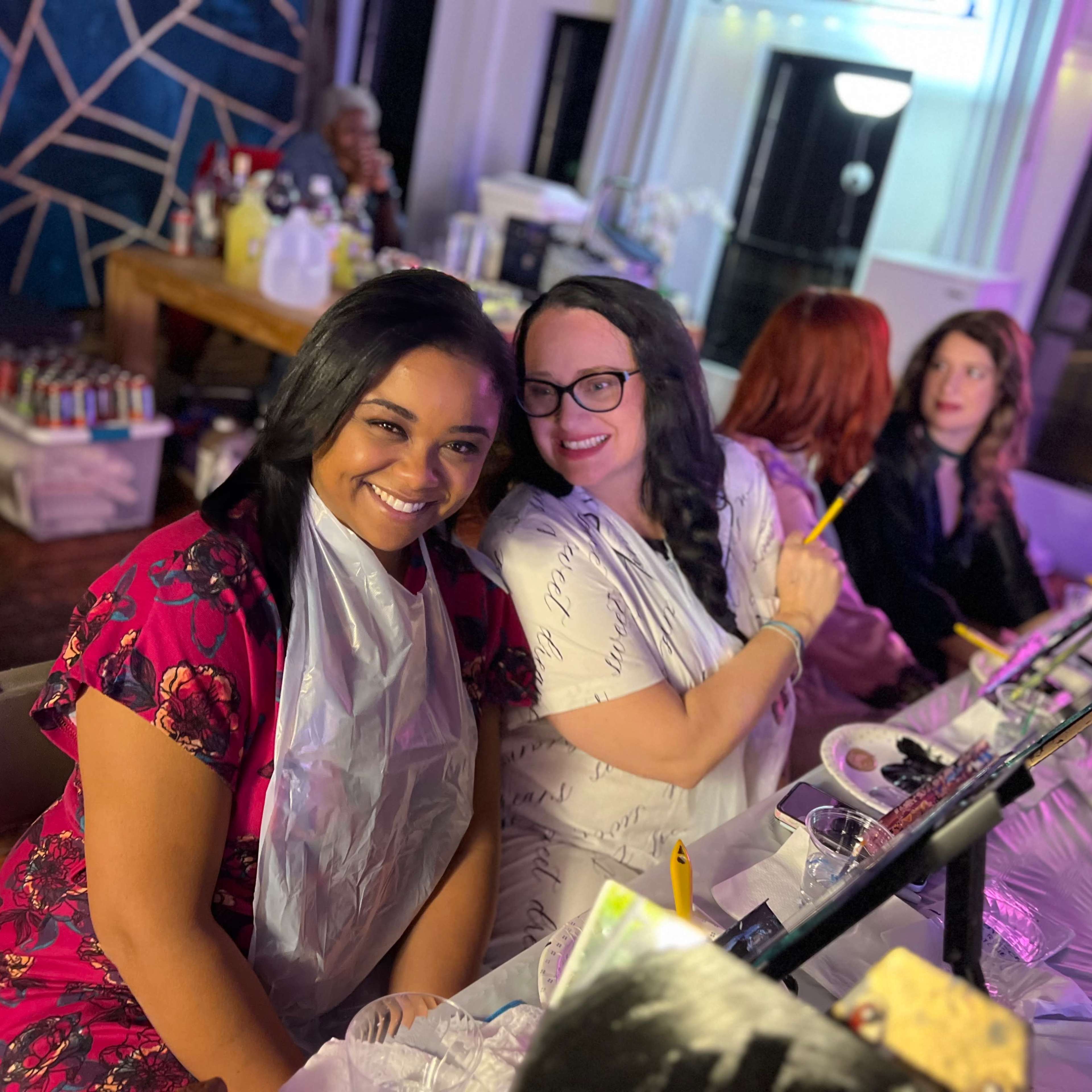 Two women sit next to each other at a table with art supplies, wearing protective bibs while painting.