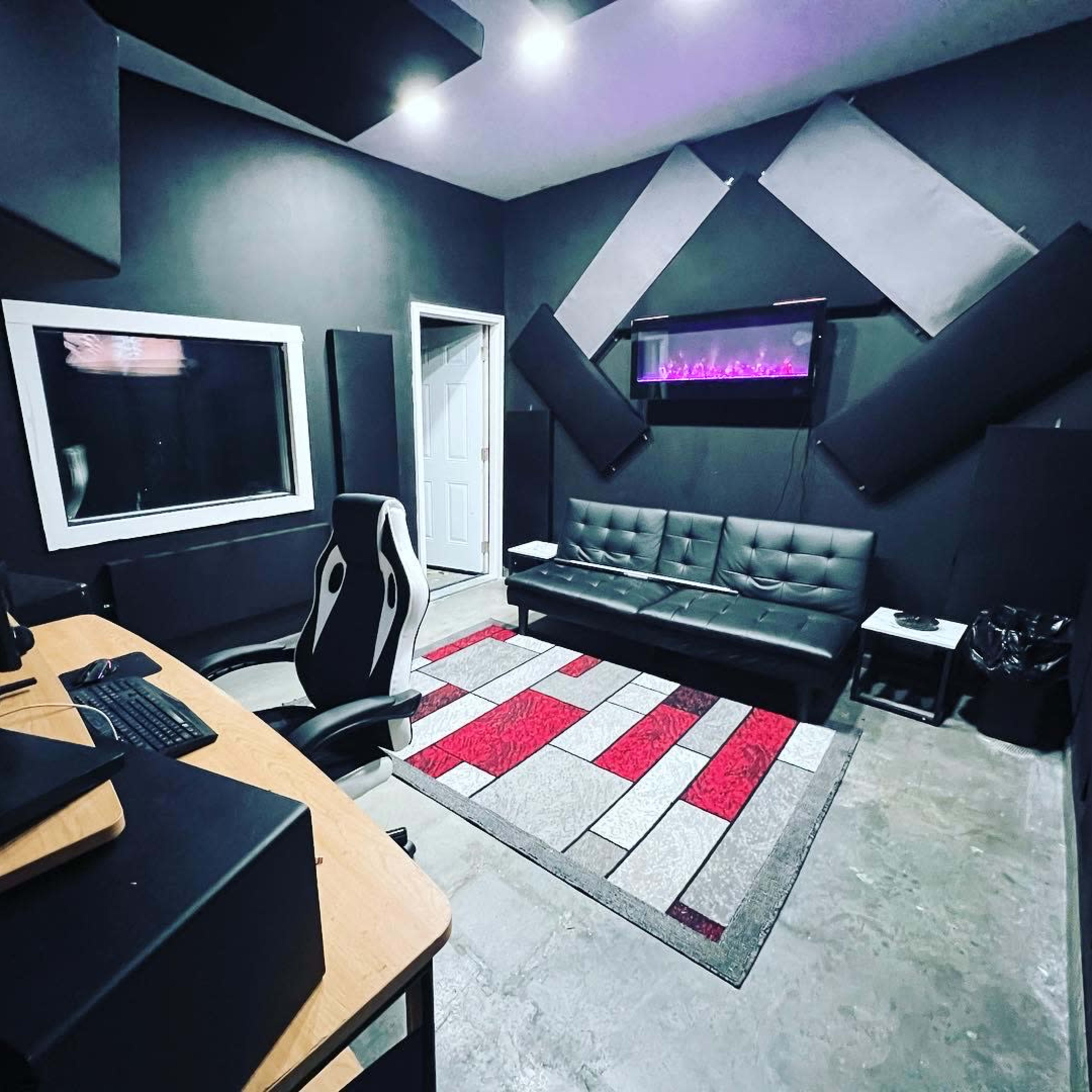 A soundproof studio room features a dark color scheme, a desk with a computer, a black couch, and an electronic display mounted on the wall.
