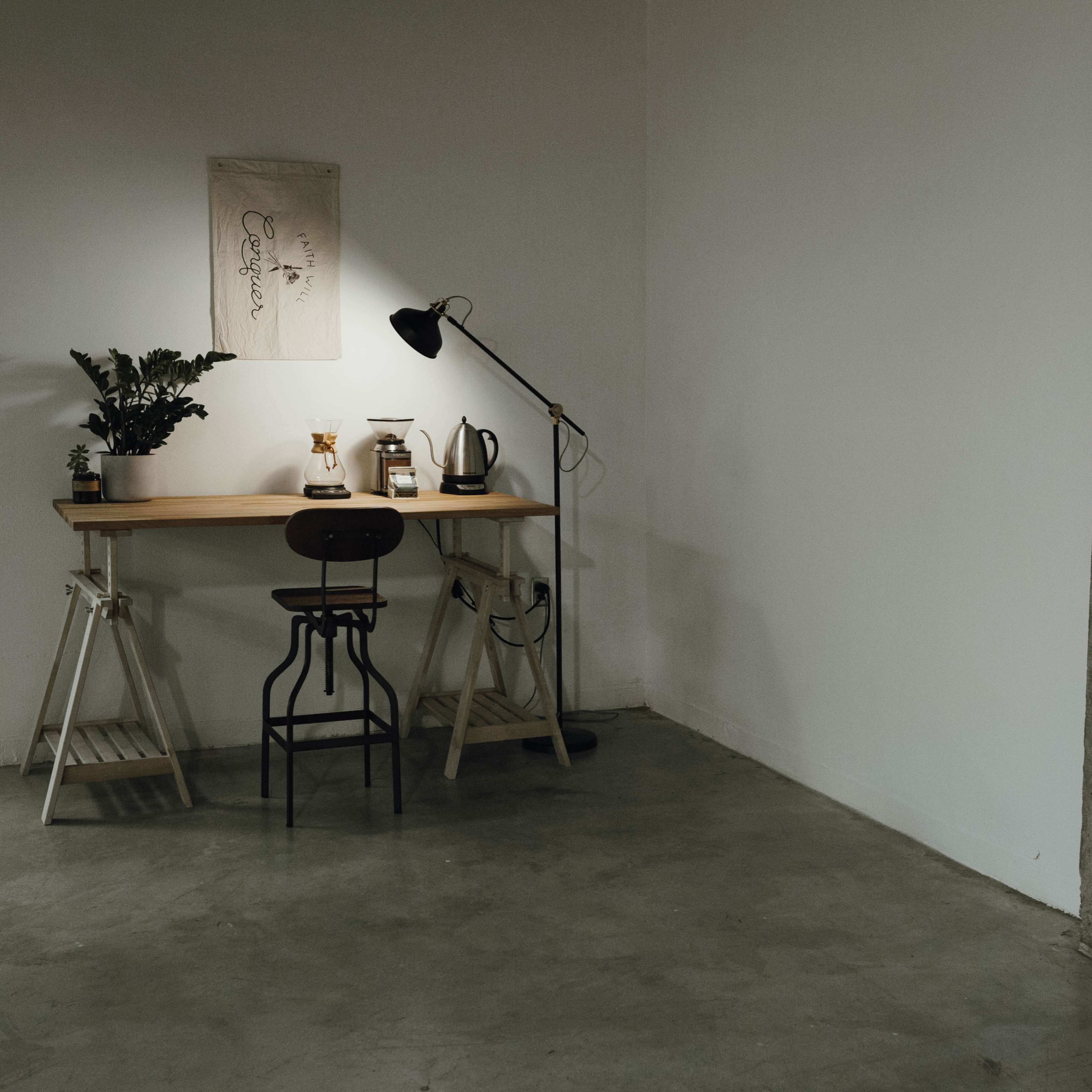 A minimalist workspace features a wooden table with a chair, a lamp, and plants against a plain wall, with a concrete surface nearby.