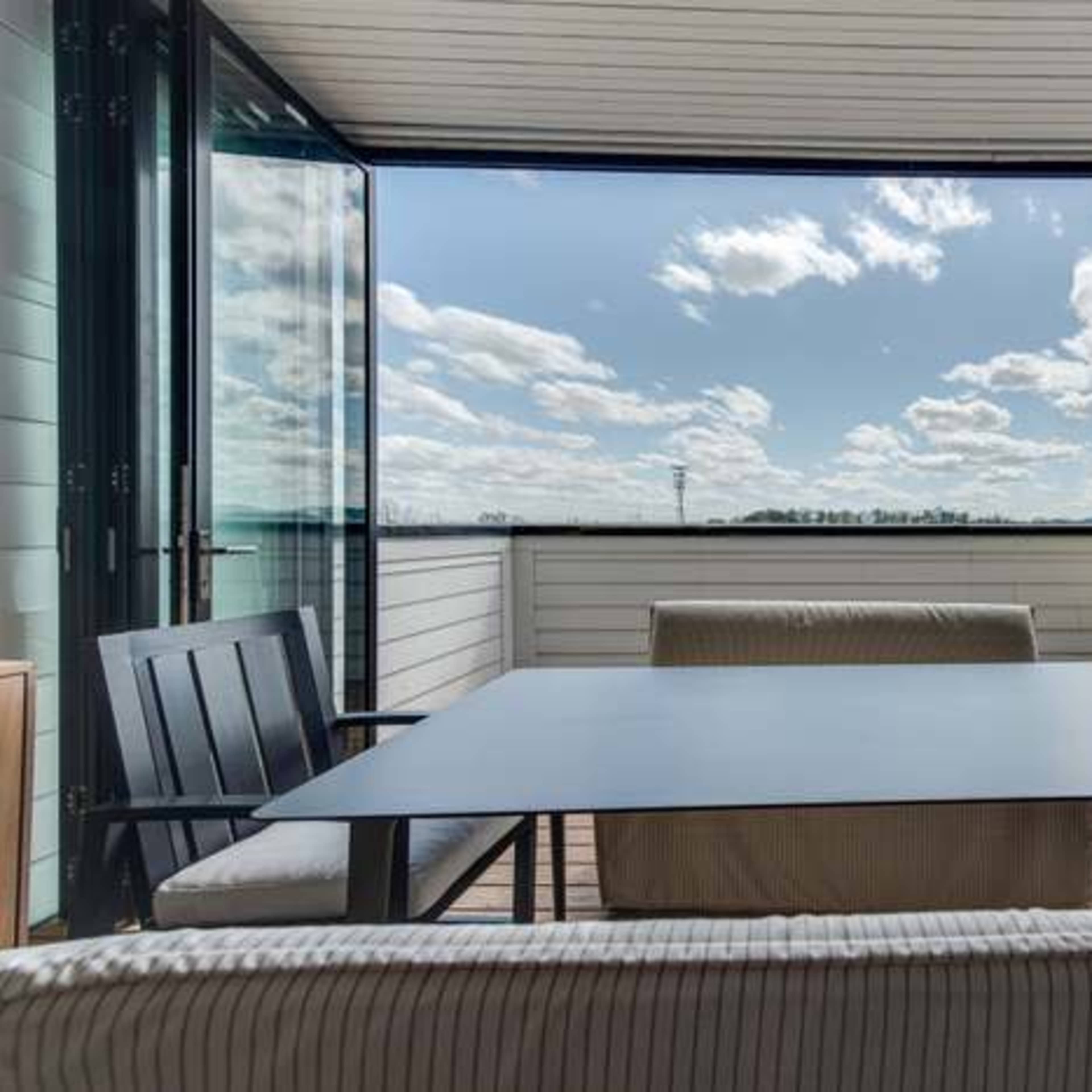 A modern outdoor patio features a table and chairs with large sliding doors opening to a view of a cloudy sky and landscape.
