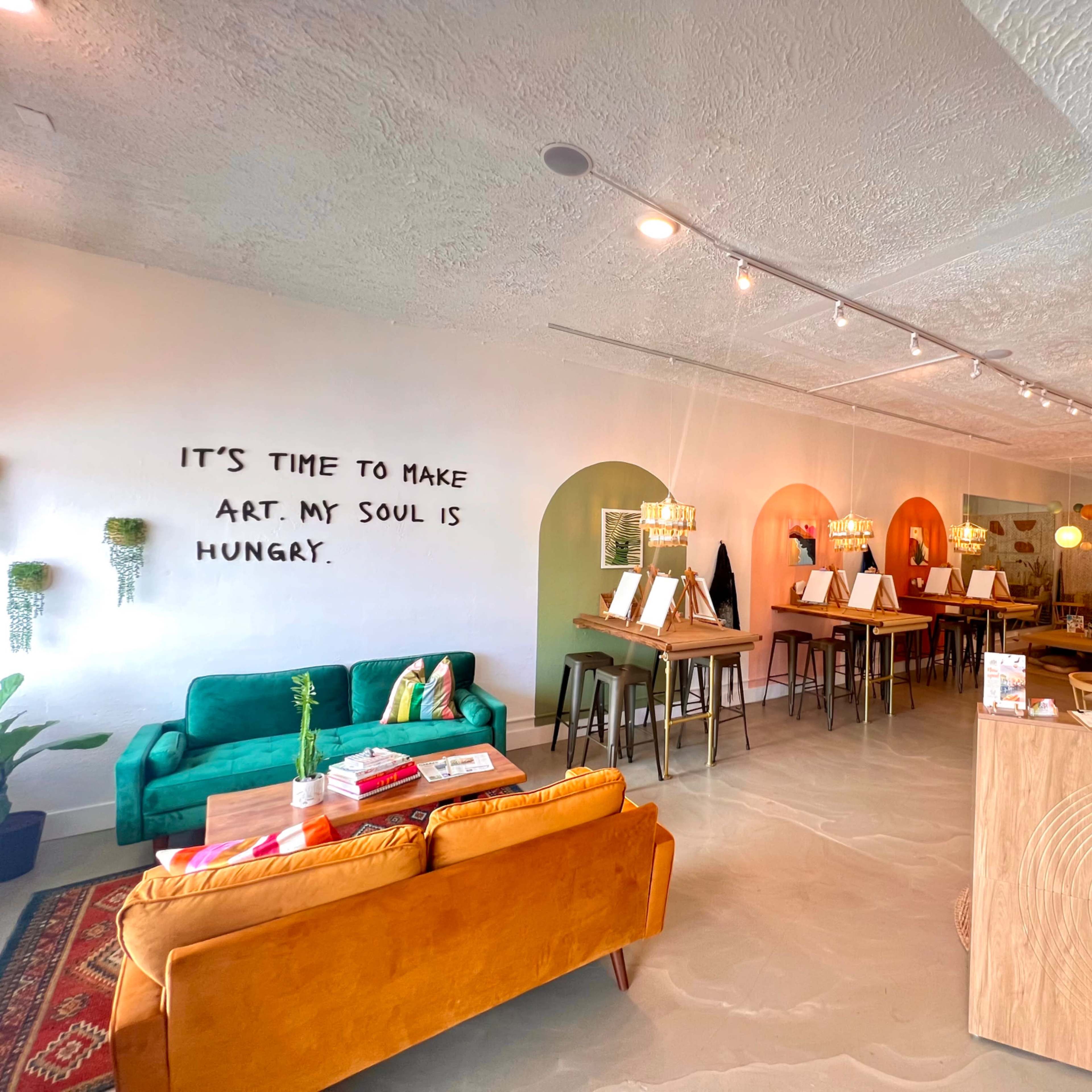 An interior space with a cozy seating area, a bar-like counter, and multiple tables set for art activities, featuring vibrant decor and text on the wall.