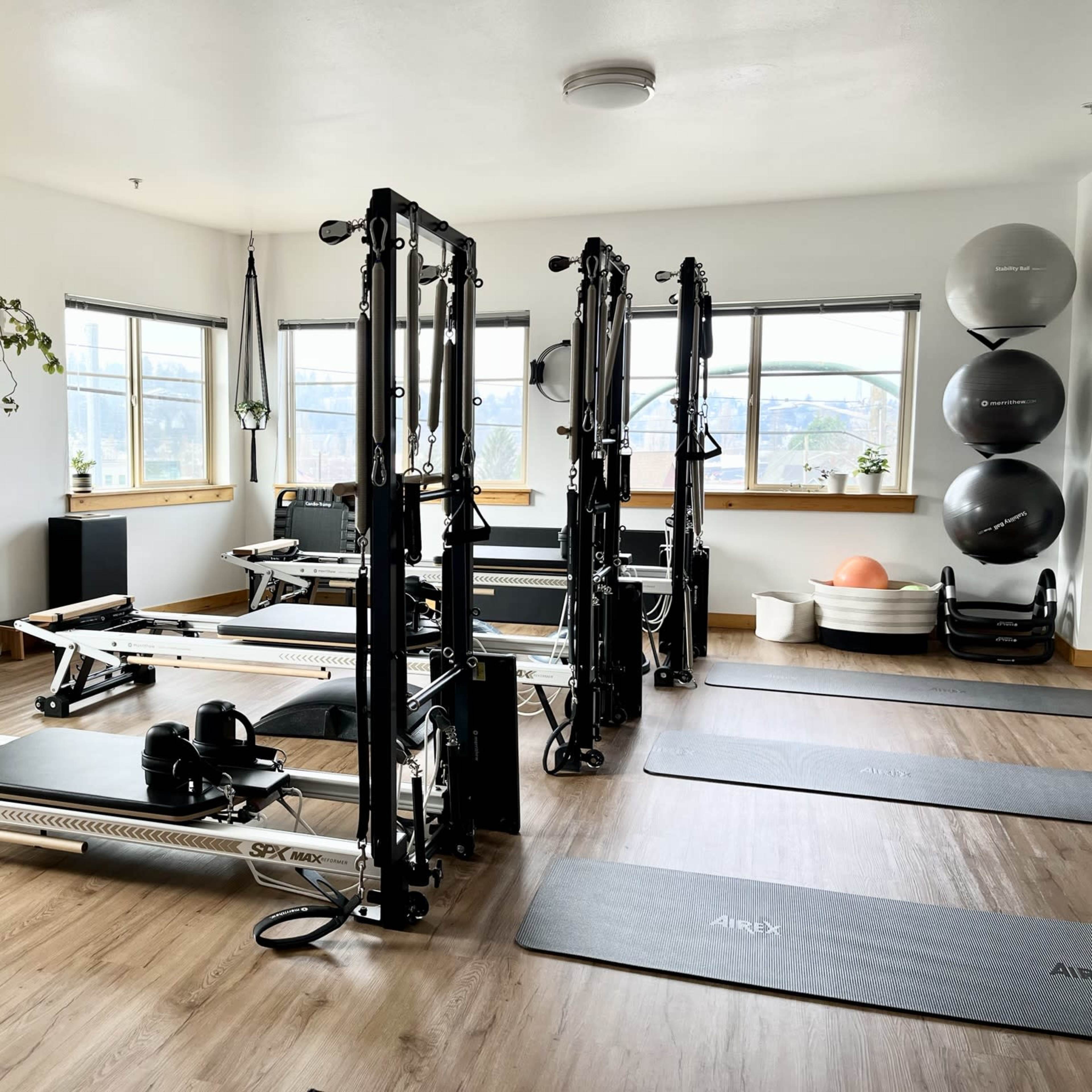 A well-lit fitness studio features reformer machines, exercise mats, and various workout equipment arranged neatly on a wooden floor.