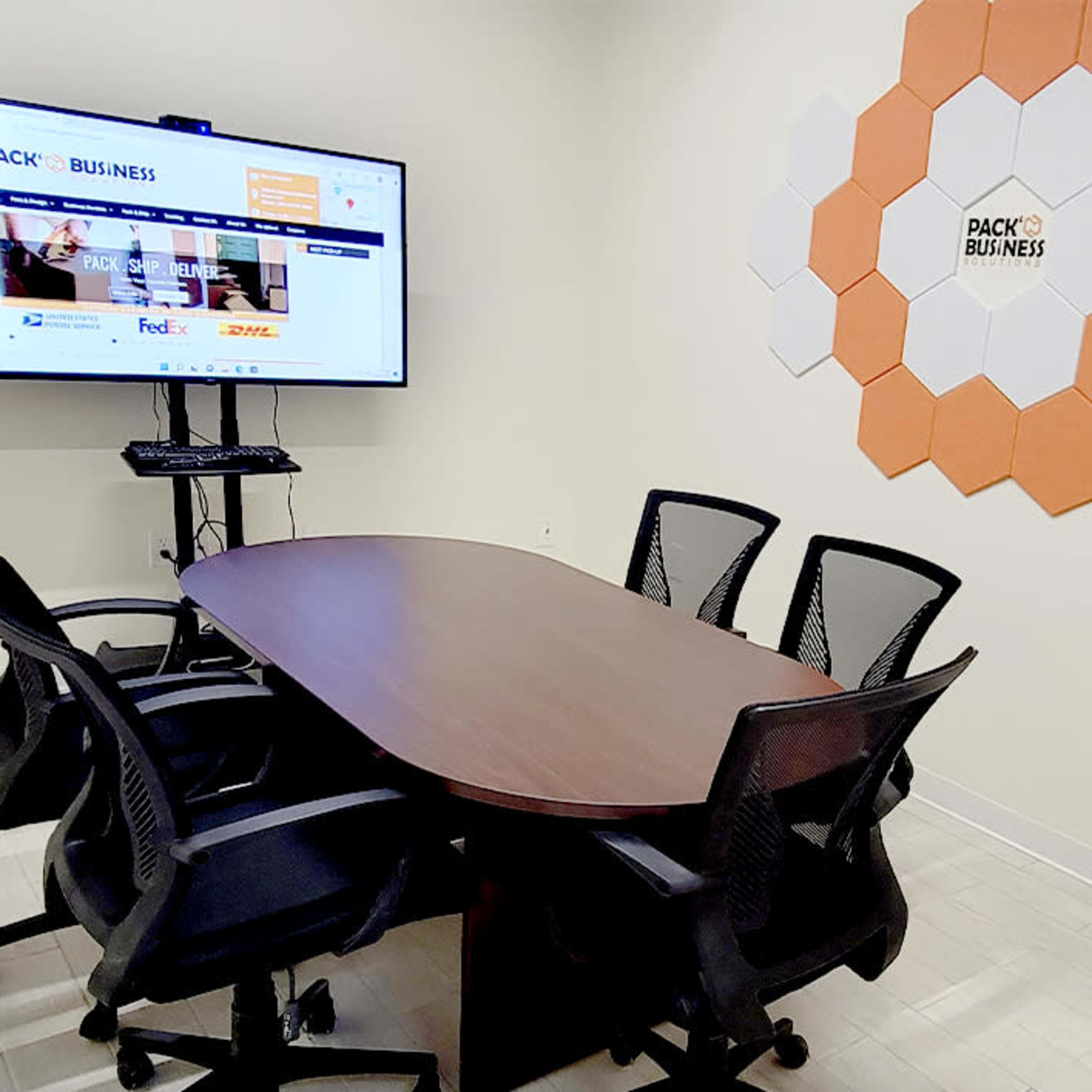 A meeting room with a rectangular wooden table, six black mesh chairs, a large screen displaying a website, and a wall decorated with hexagonal orange and white panels.