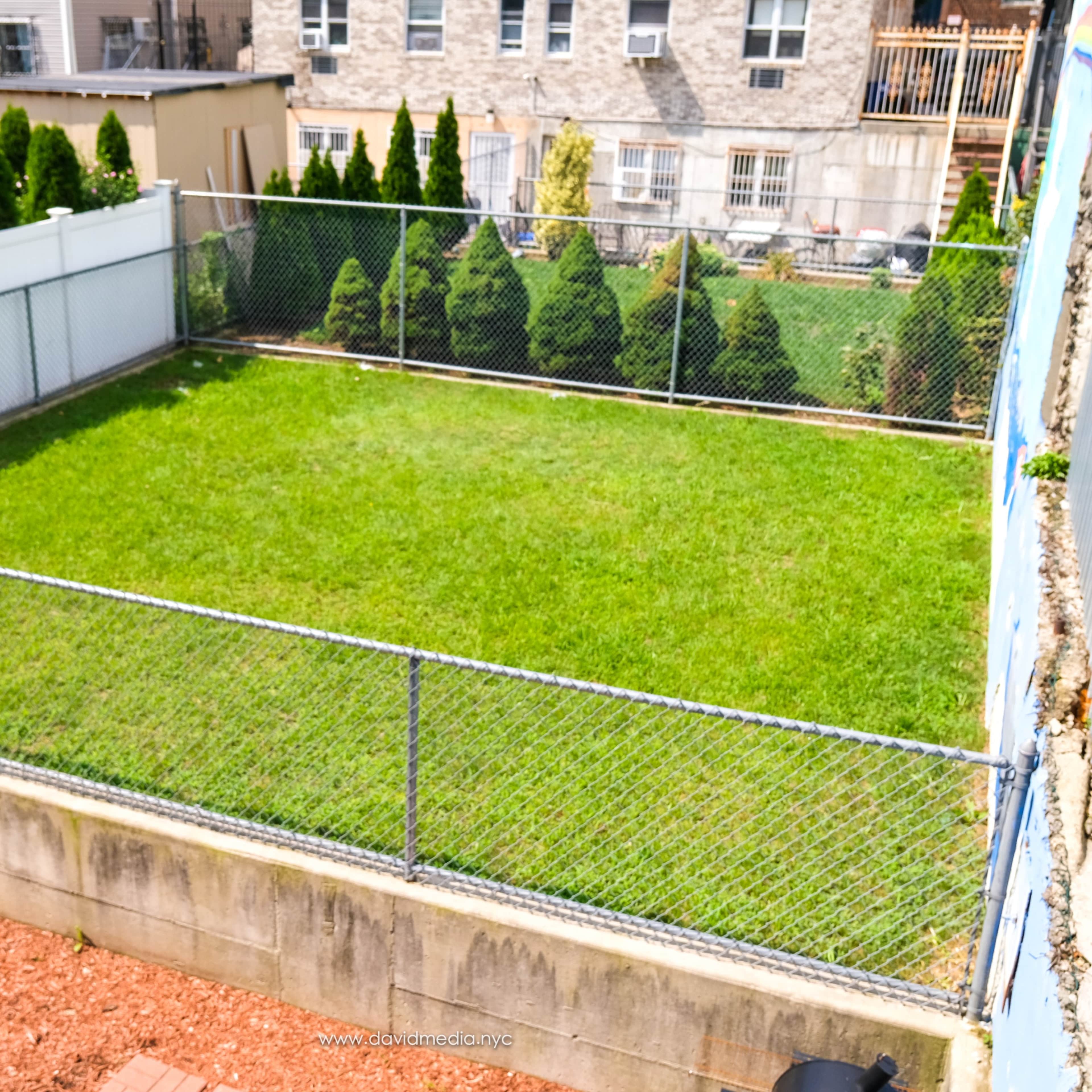 A fenced grassy yard with a concrete base and a view of neighboring buildings in the background.