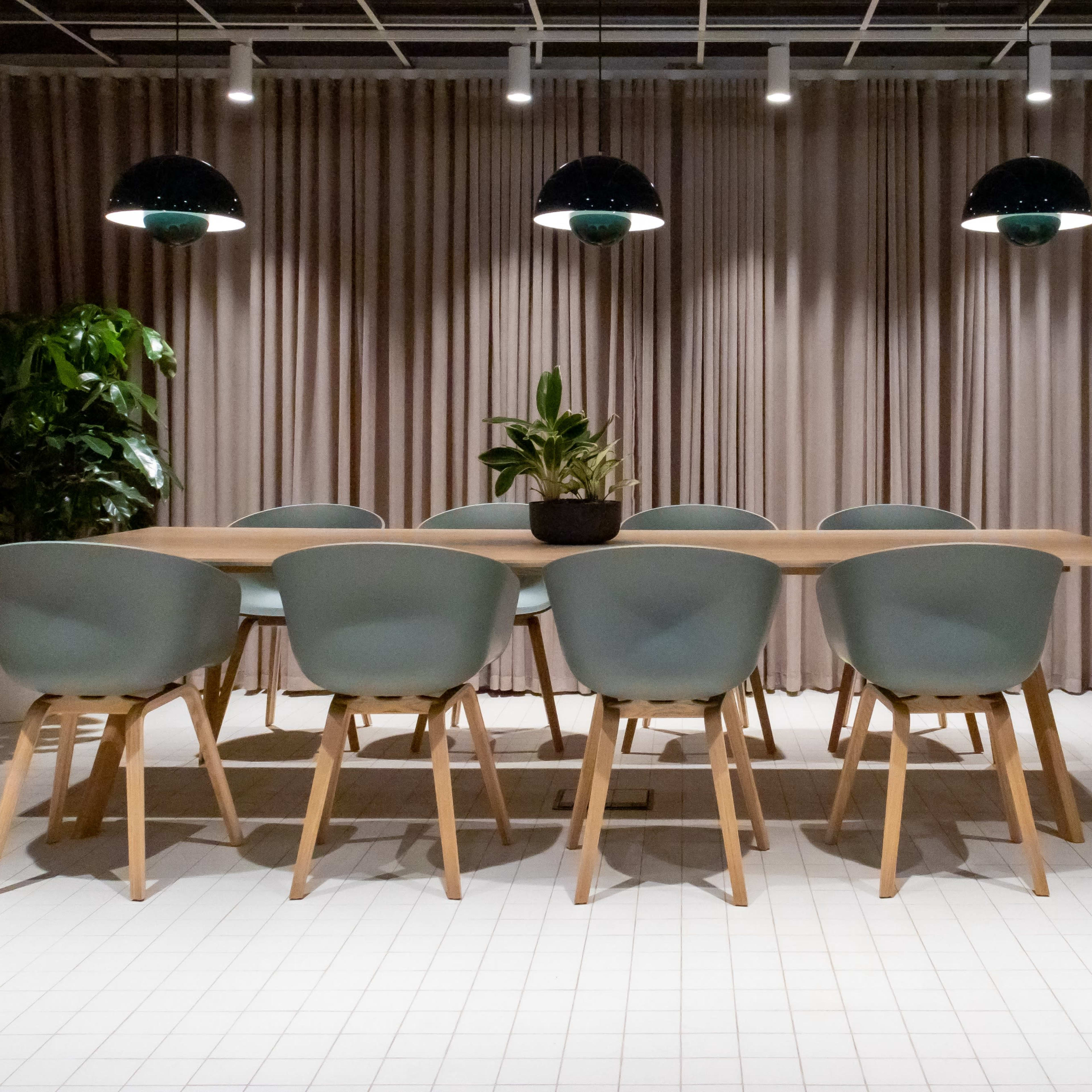 A long wooden dining table with light blue chairs is situated under black pendant lights in a room with soft beige curtains and indoor plants.