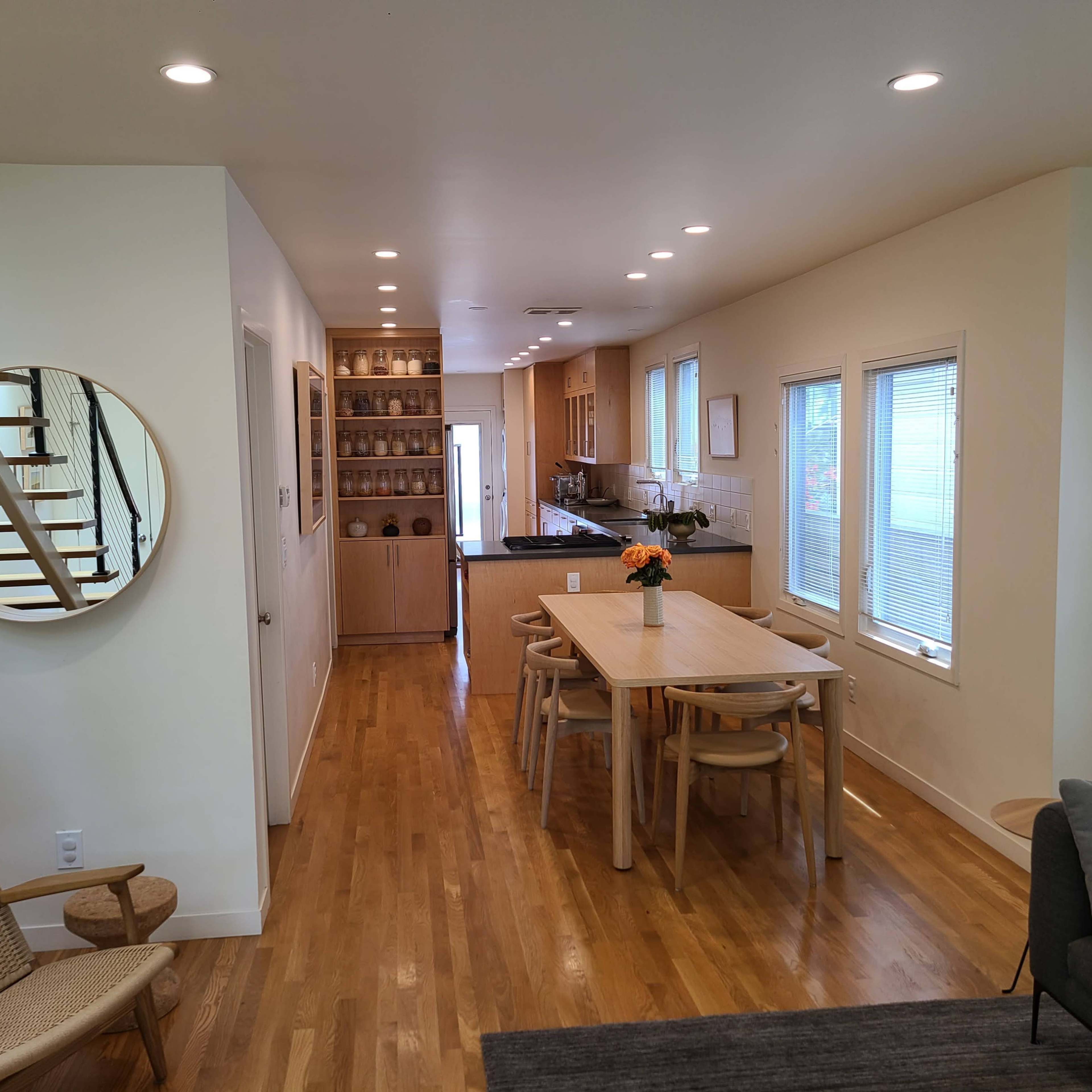 A modern interior space features a dining area with a wooden table and chairs, a kitchen in the background, and a staircase leading to the upper level.