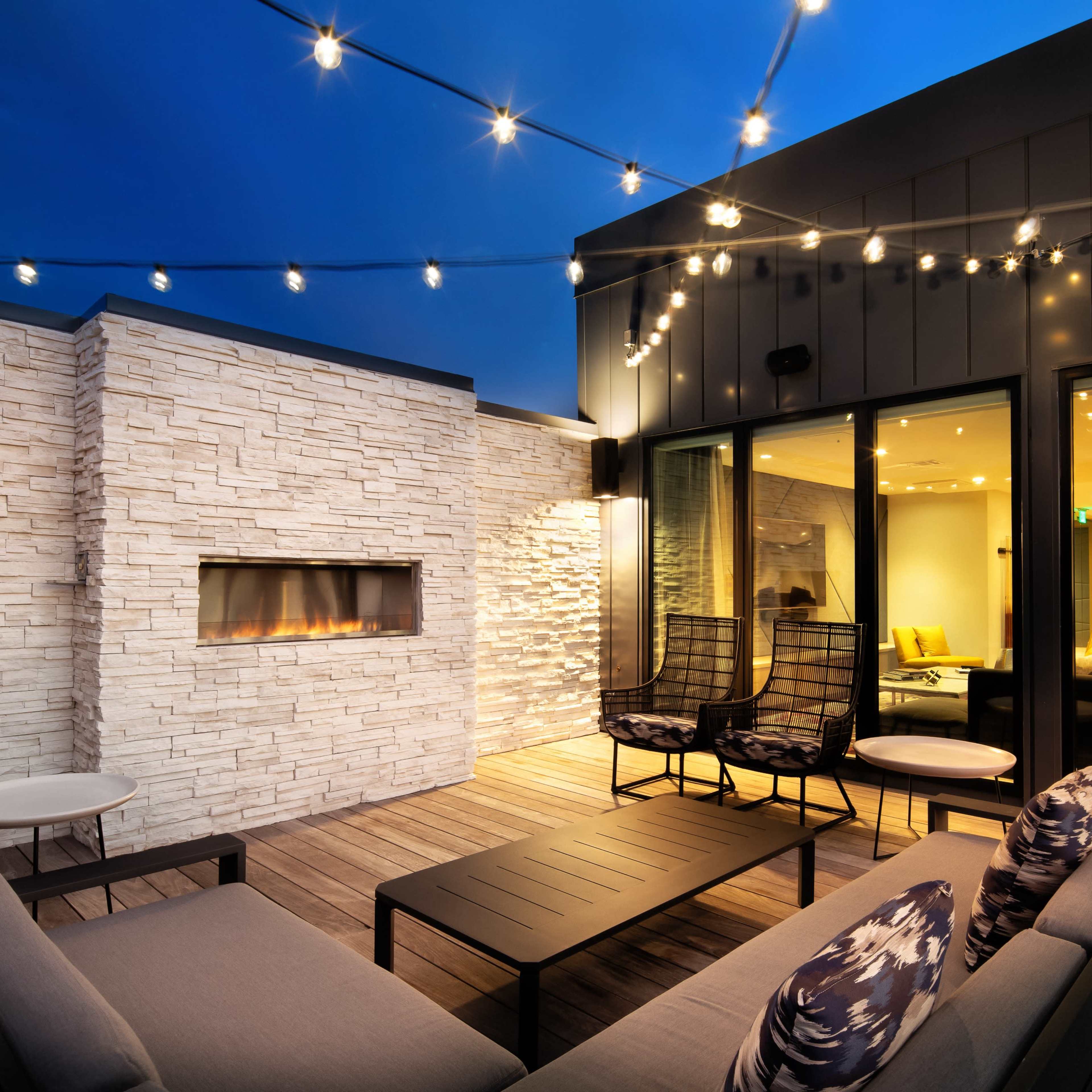 A modern outdoor seating area features a stone wall with a fireplace, surrounded by string lights, and is furnished with a sofa and chairs on a wooden deck.