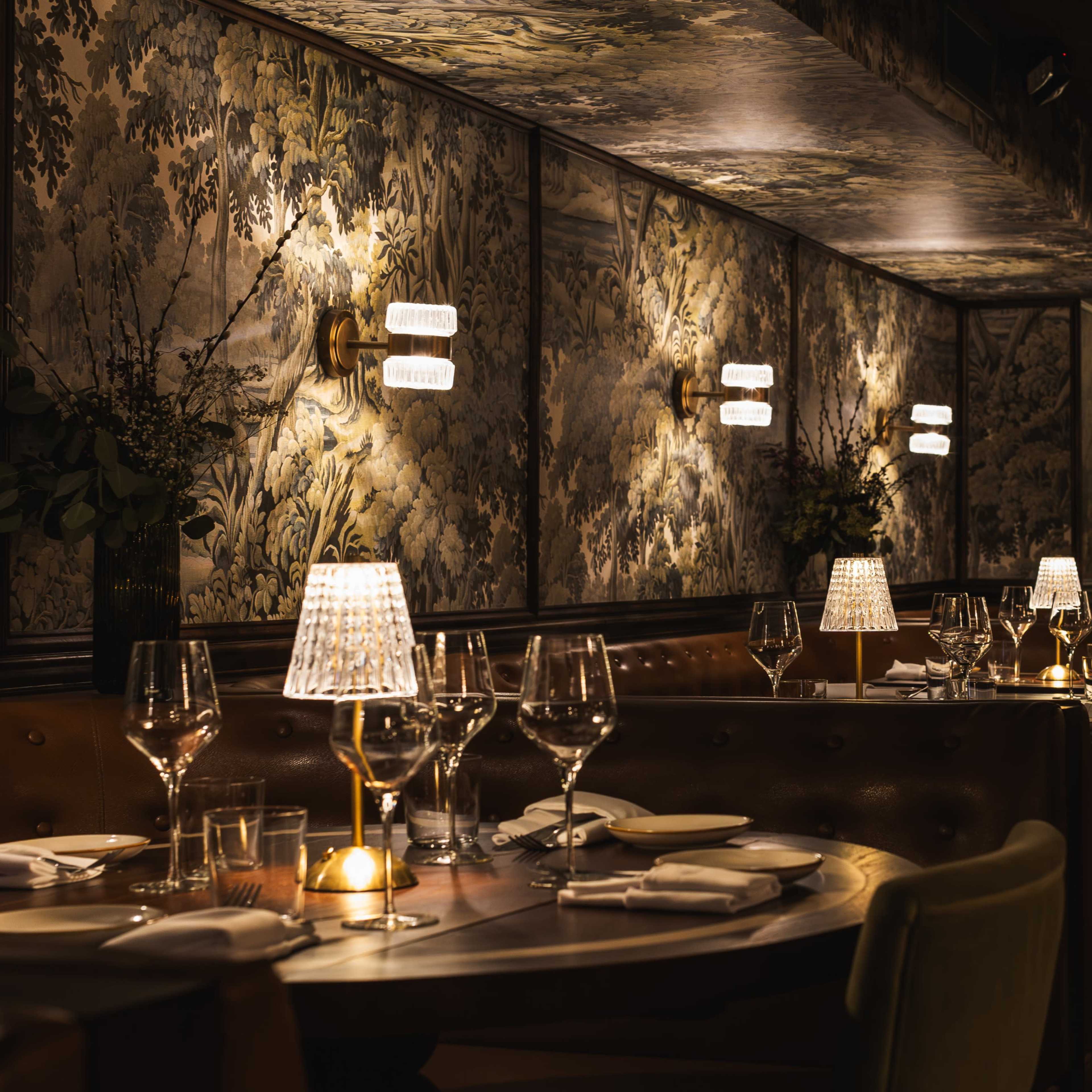 A dimly lit restaurant featuring elegant table settings with glassware and lamps, surrounded by ornate wallpaper and decorative wall sconces.