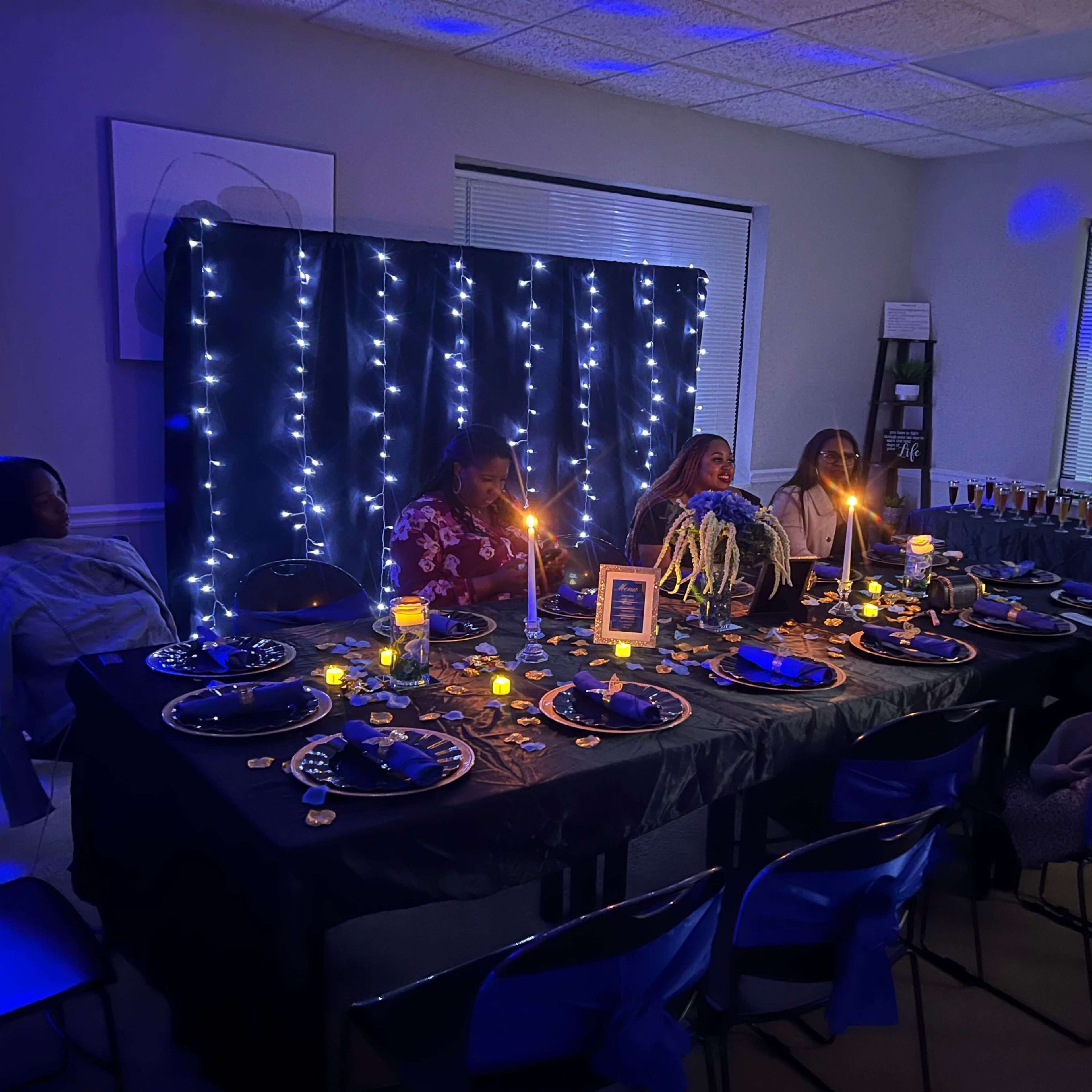 A dimly lit room features a dinner table set for five, adorned with candles and blue fairy lights in the background.