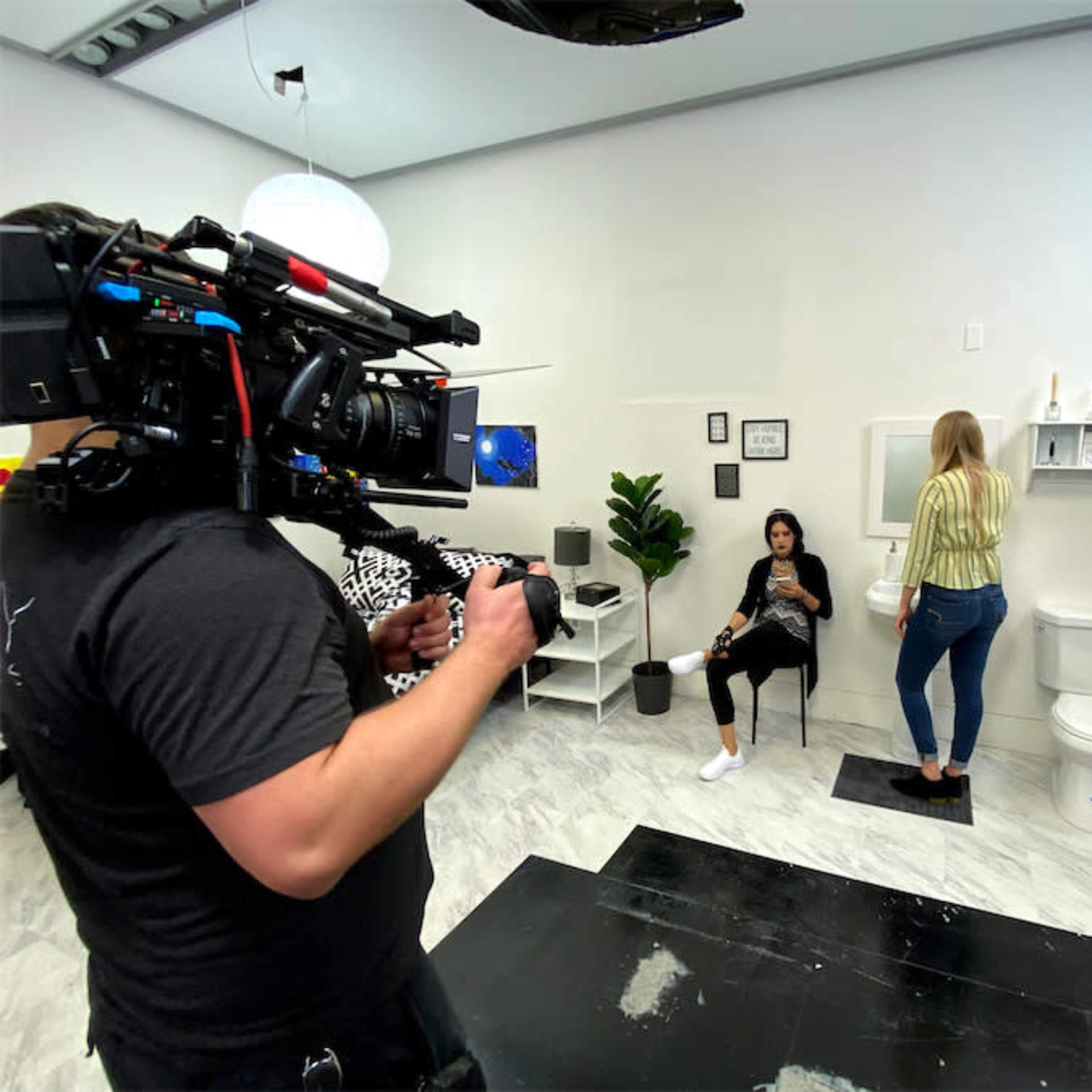 A camera crew shoots a scene in a bathroom set where one woman is sitting while another stands near the sink.