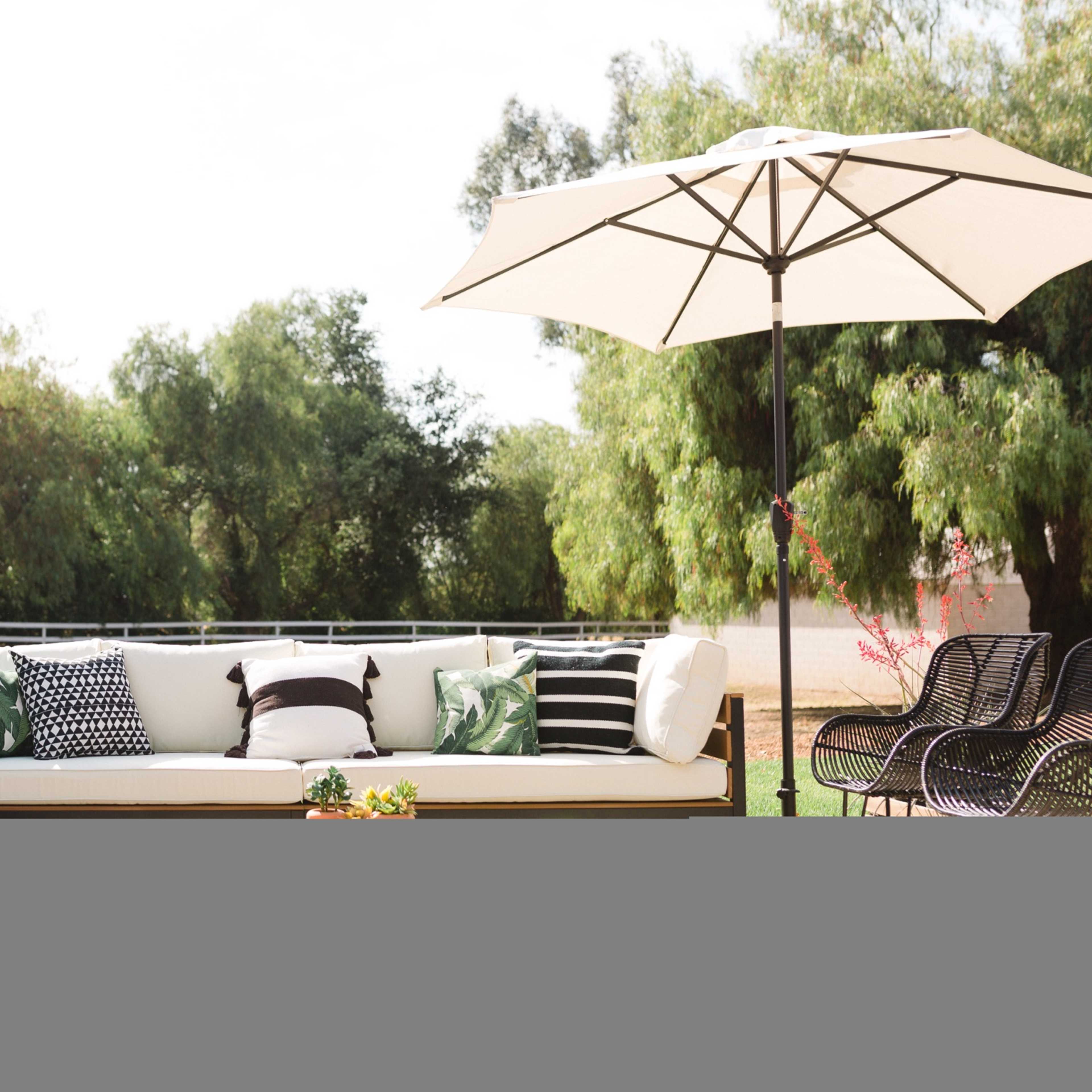 A patio area features a large sofa with various patterned cushions, surrounded by greenery and a large umbrella providing shade.
