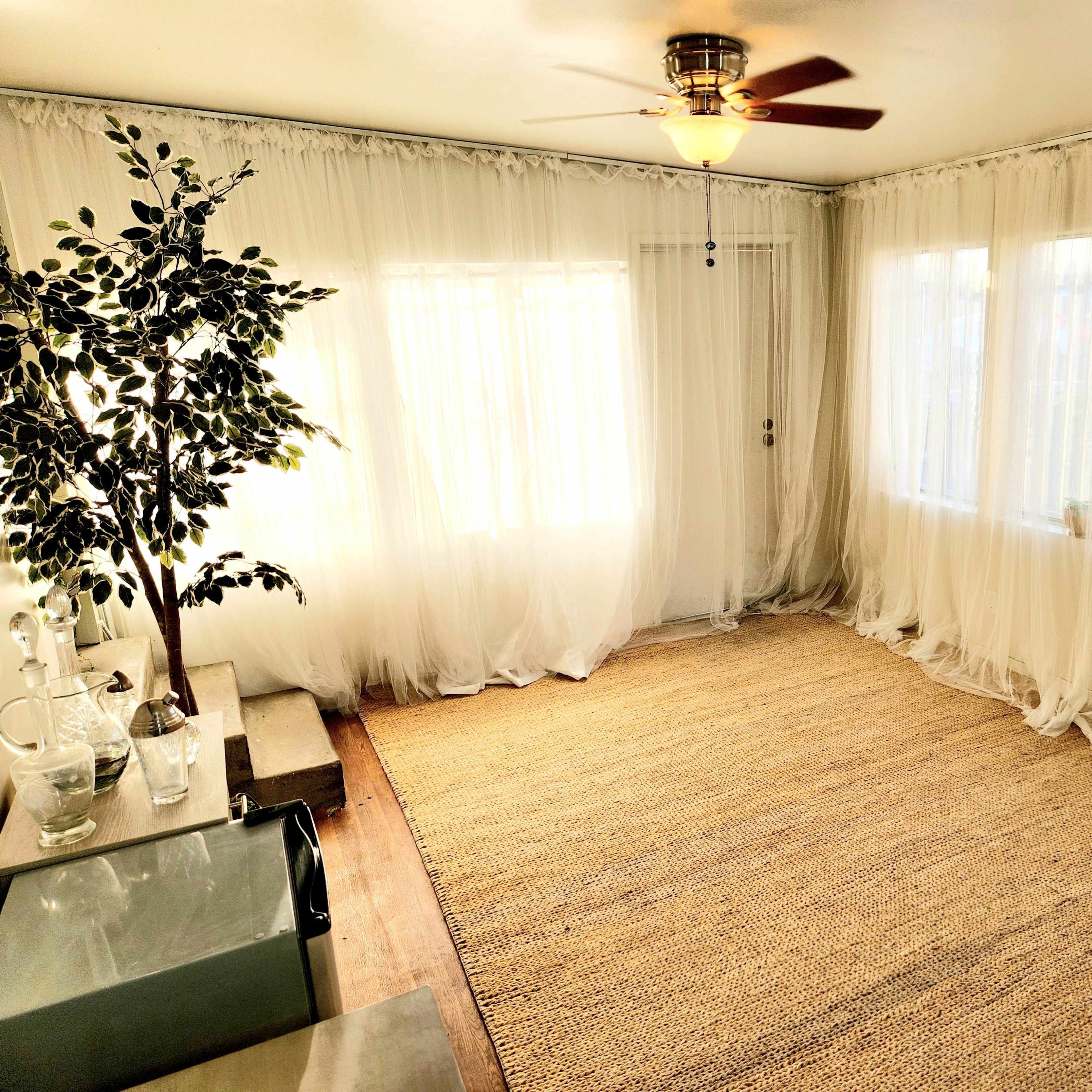 A sunlit room features sheer white curtains, a ceiling fan, a woven rug, and a potted plant in the corner.